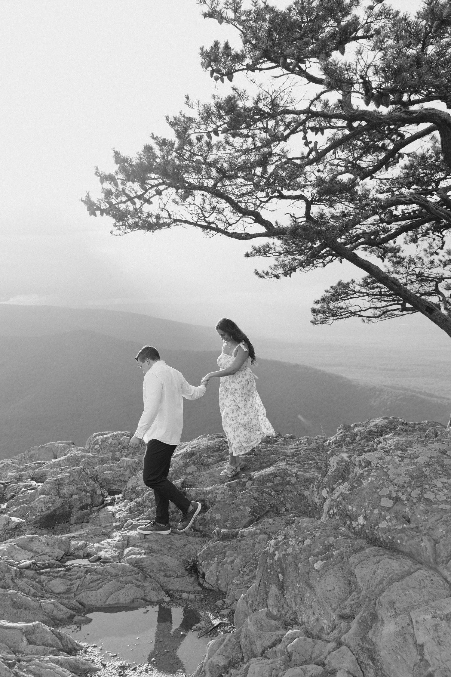 Black and white photo of couple holding hands walking on rocky mountain overlook.