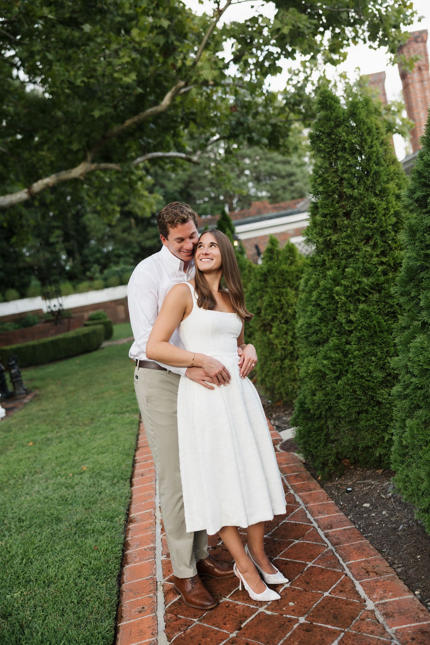 Couple embracing in a garden, woman in white midi dress and heels, man in white shirt and khakis, surrounded by greenery.