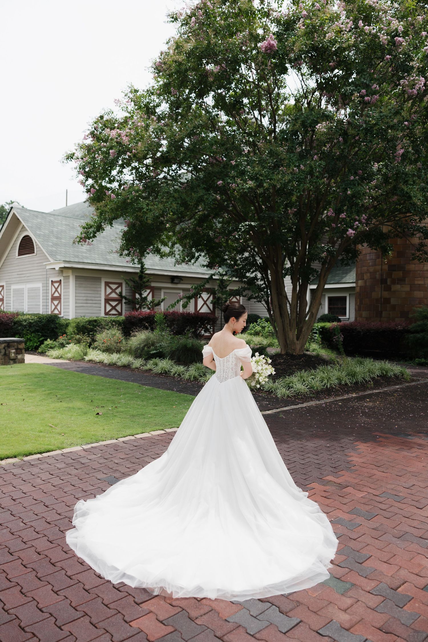 Bride in elegant white off-shoulder gown with long train stands on brick path, holding bouquet before lush garden backdrop.