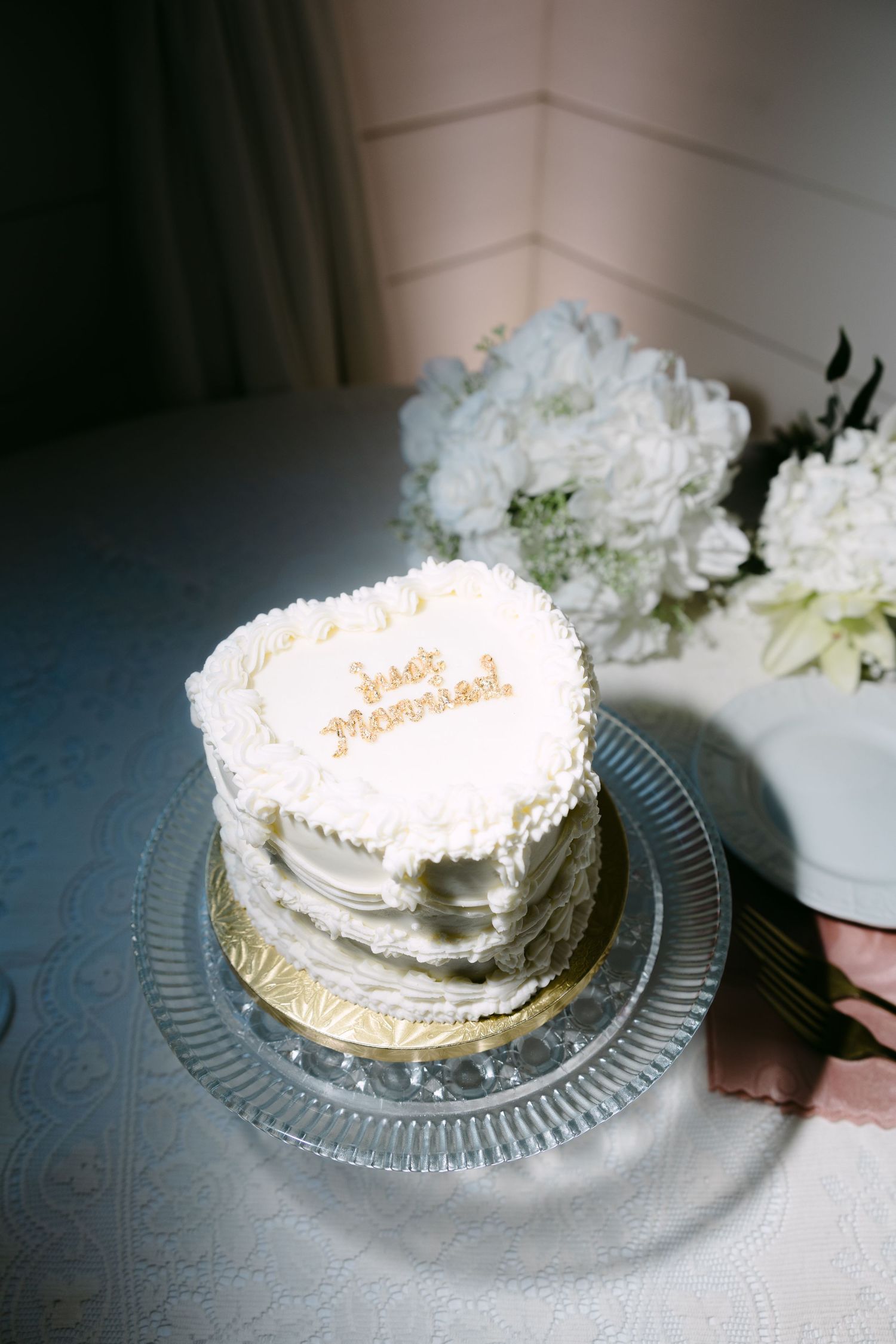 Heart-shaped white wedding cake reading Just Married on glass plate with white floral arrangement.