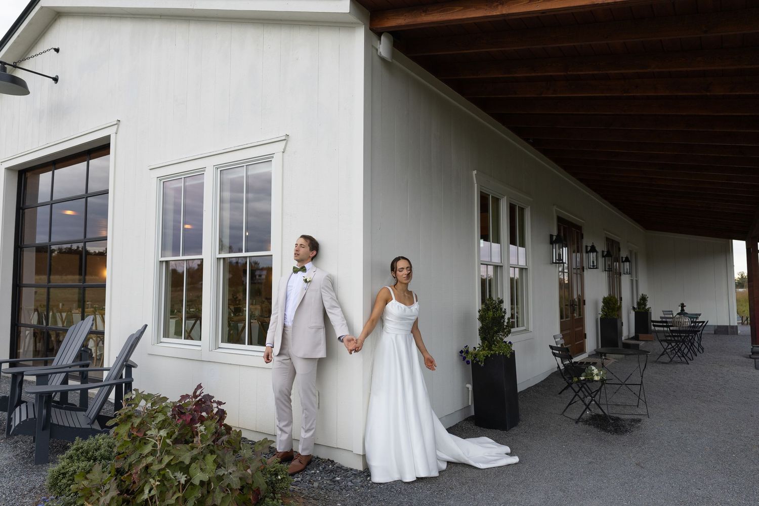 Bride and groom holding hands outside a white farmhouse wedding venue with covered porch and string lights.