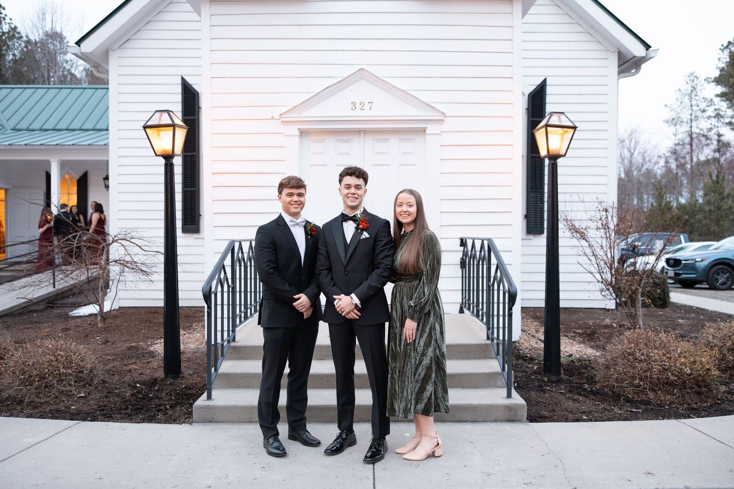 Three young adults in formal attire posing before a white chapel entrance.