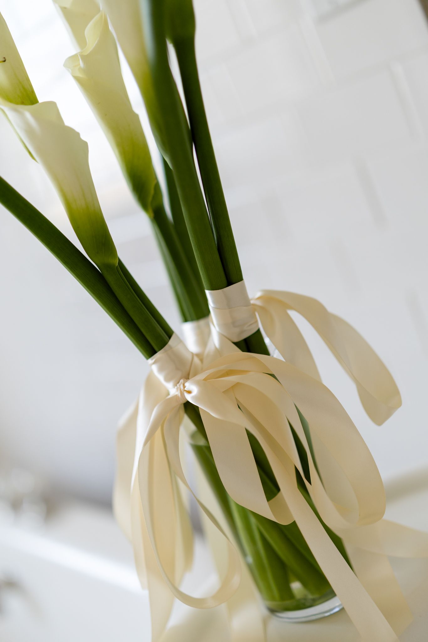 Elegant white calla lily bridal bouquet tied with ivory satin ribbon in clear glass vase.