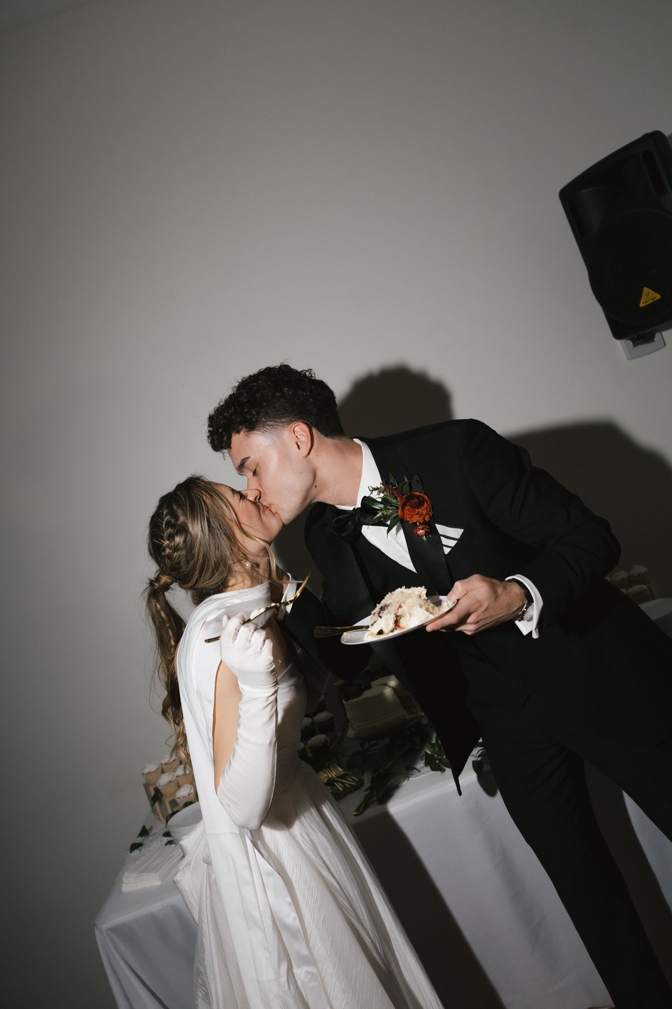 Bride and groom sharing a romantic kiss while holding a plate of wedding cake.