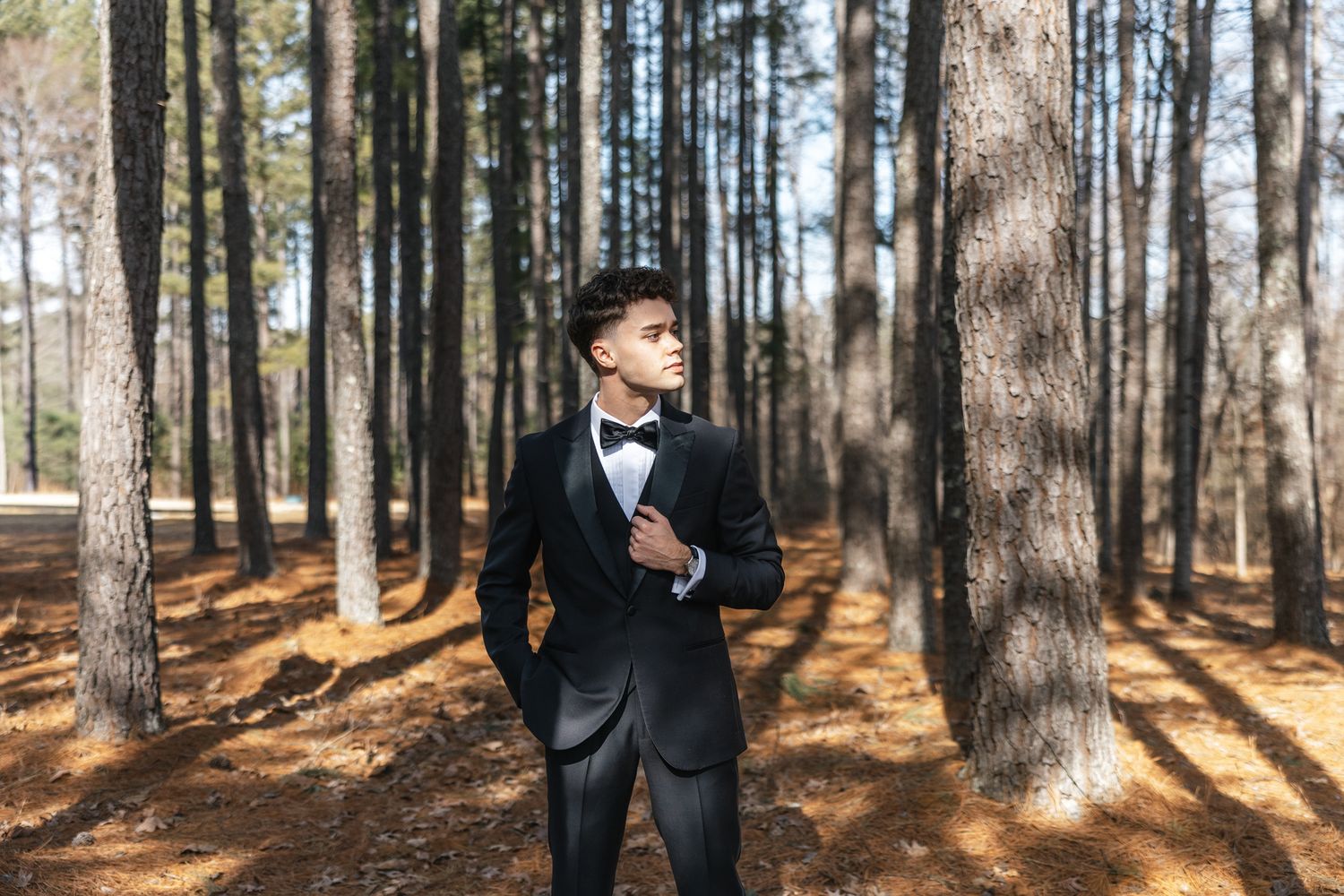 Groom in black tuxedo with bow tie standing among tall pine trees outdoors.