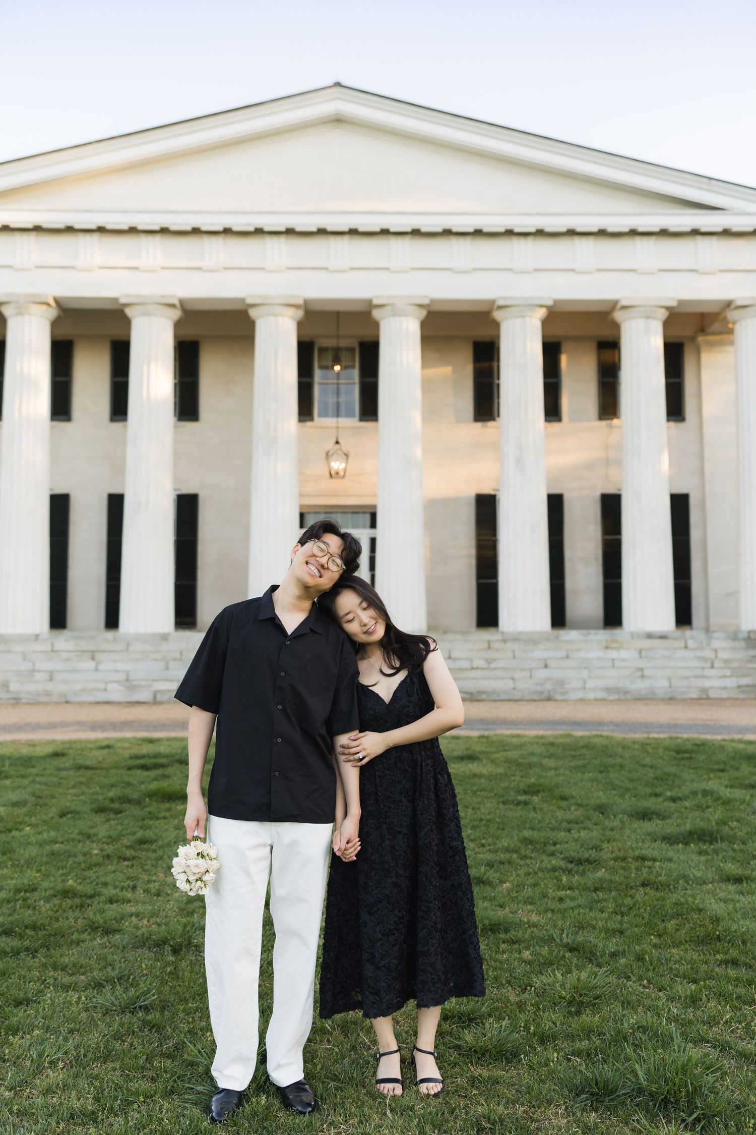 A couple embraces on a green lawn in front of a white neoclassical building at dusk.