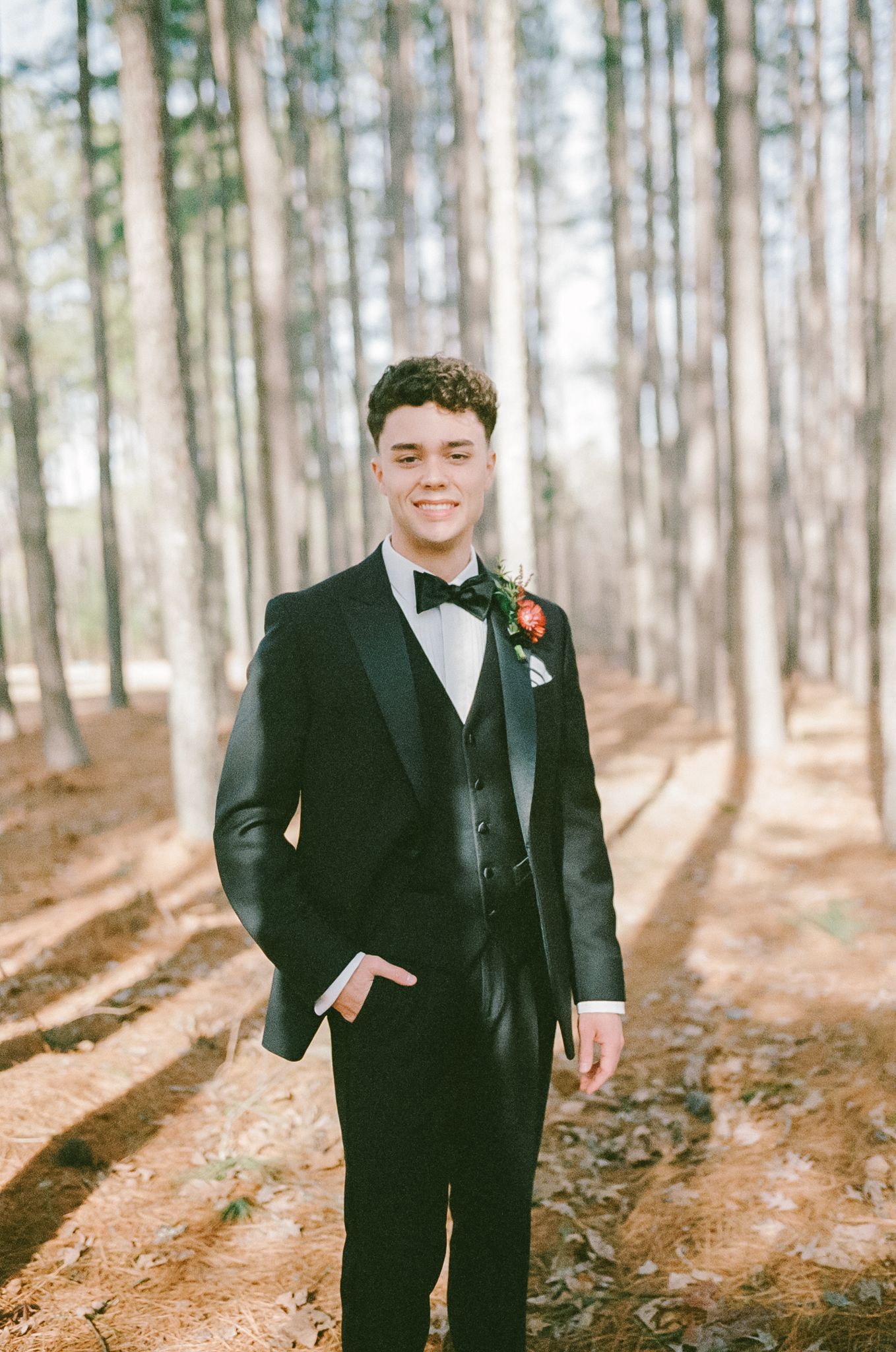 Young groomsman in black tuxedo with boutonniere standing in sunlit pine forest photographed on 35mm film.