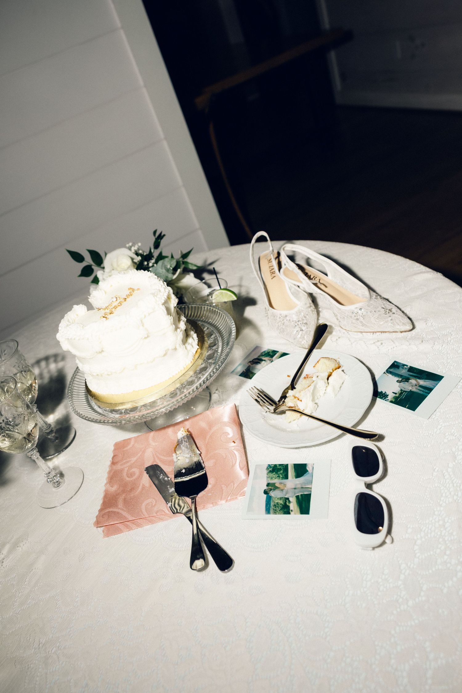 Elegant two-tier white wedding cake displayed with bridal heels and Polaroid photos on table.