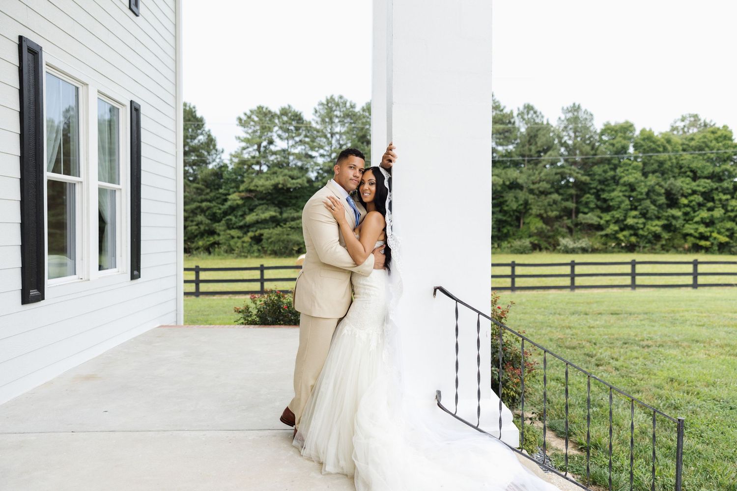 A couple in wedding attire embraces on a white farmhouse porch with a scenic green countryside backdrop.
