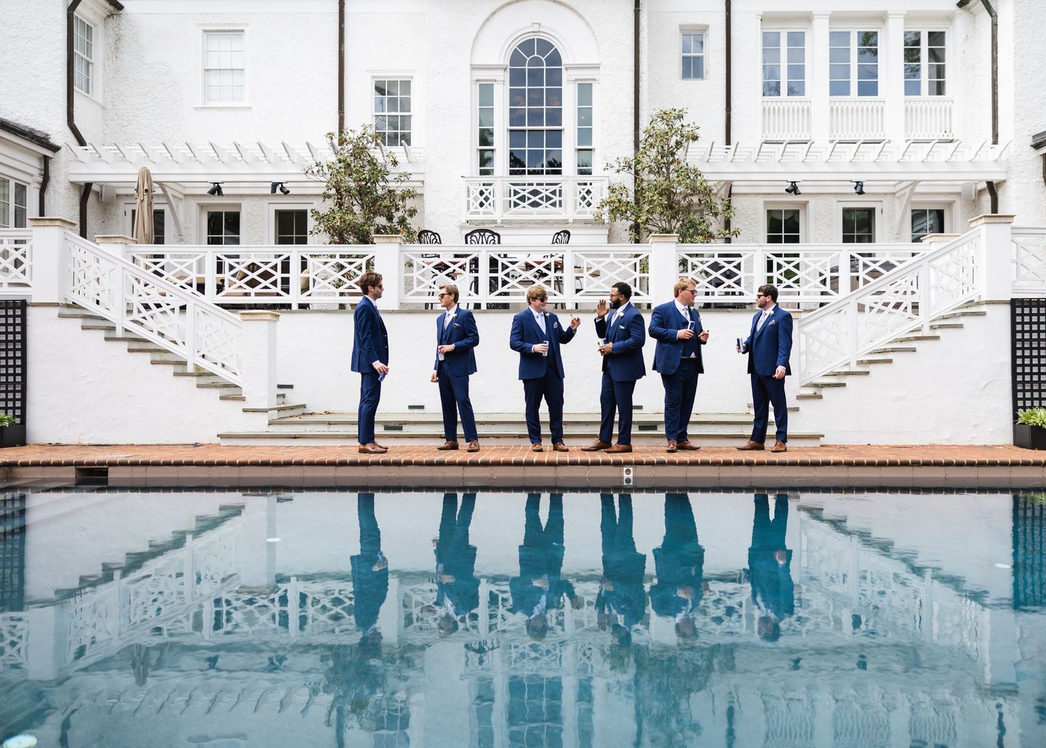 Six groomsmen in navy suits posed by a reflective pool outside an elegant white mansion.