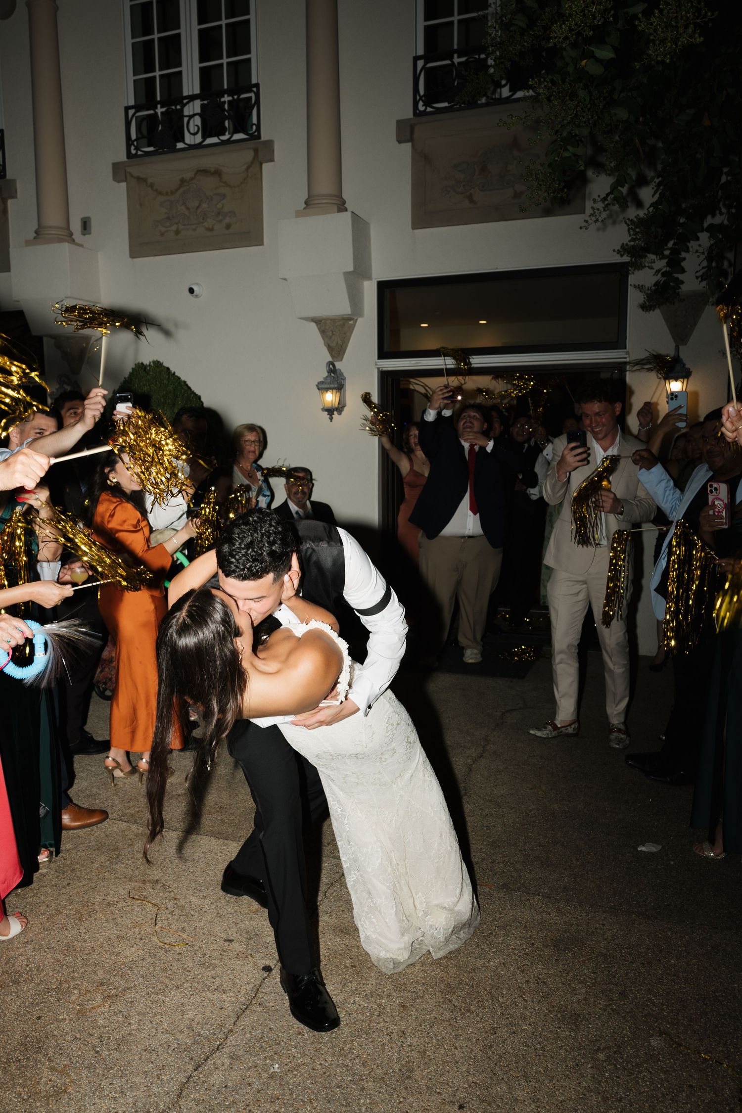Groom dips bride for a romantic kiss during wedding send-off with cheering guests and brass band outside.