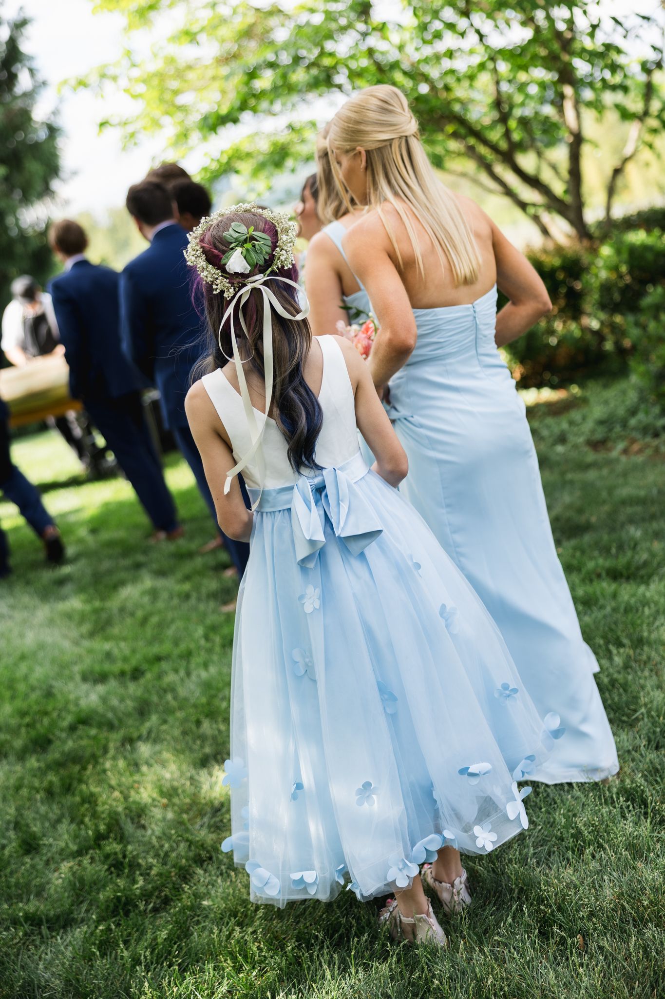 Flower girl in light blue dress with floral crown walks beside bridesmaid at outdoor wedding.