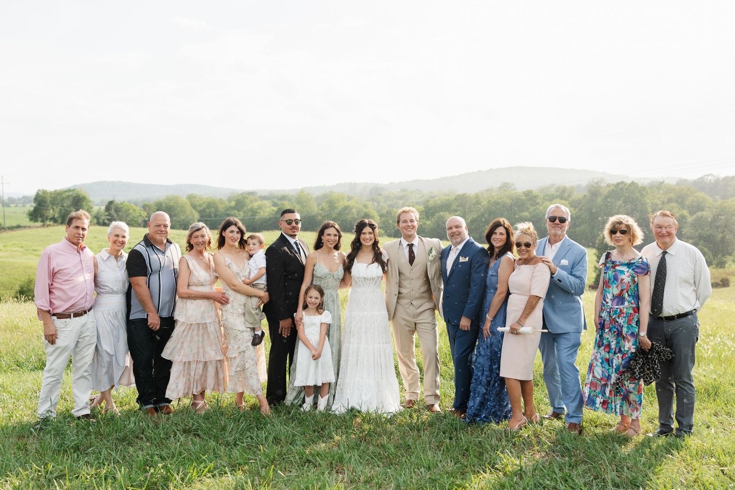 Wedding party group photo outdoors on green grass with rolling hills, bride in white gown surrounded by family.