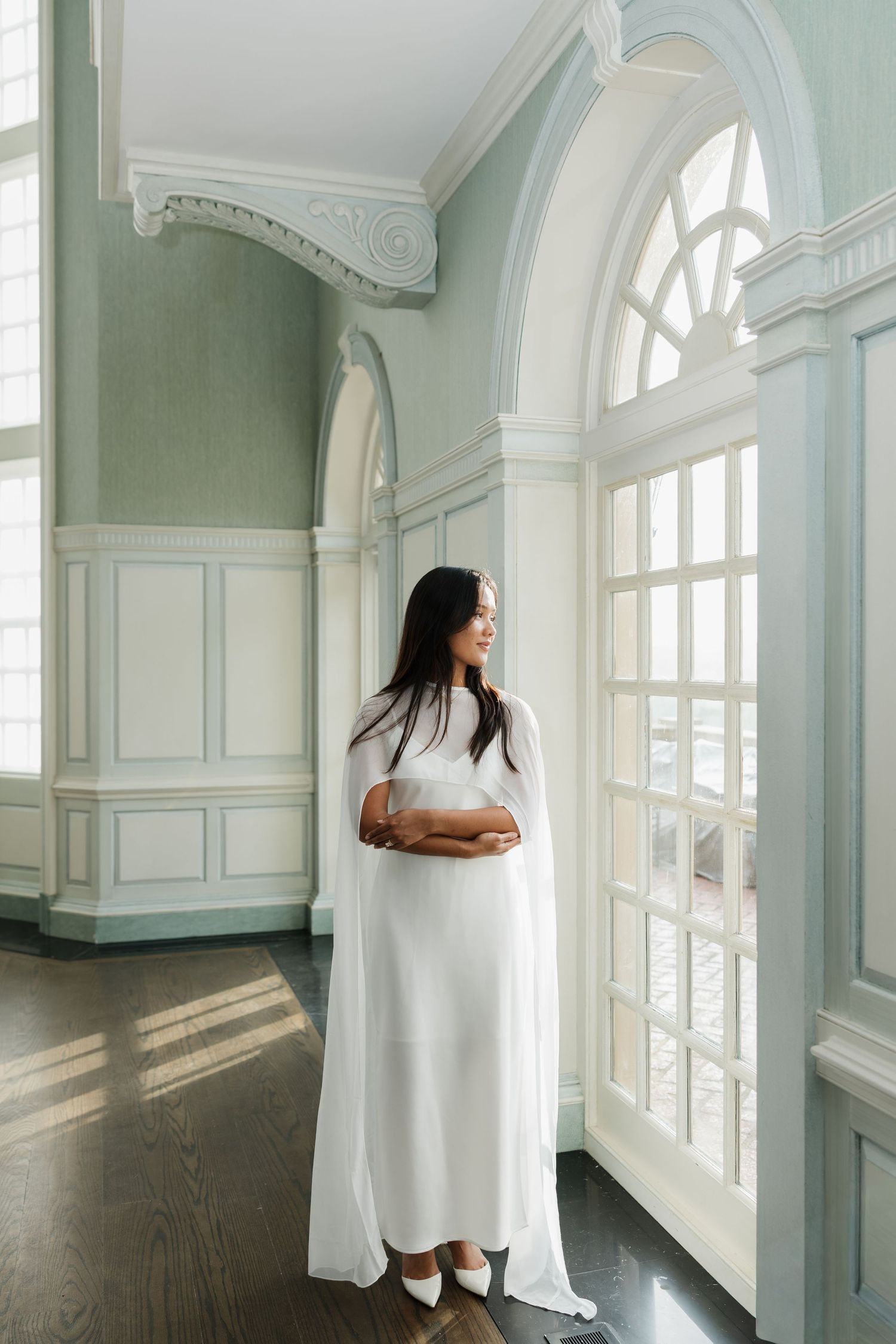 Pregnant woman in white dress standing by arched windows in elegant room with sage green walls.