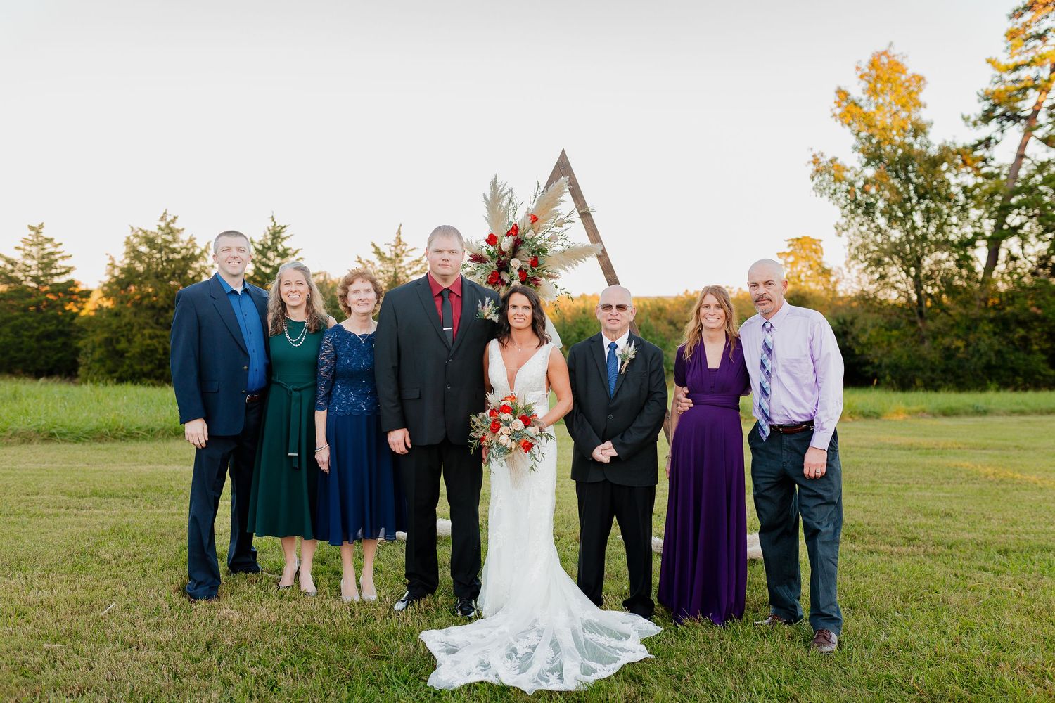 Wedding party of eight poses at golden hour beside a wooden arch on a green rural lawn.