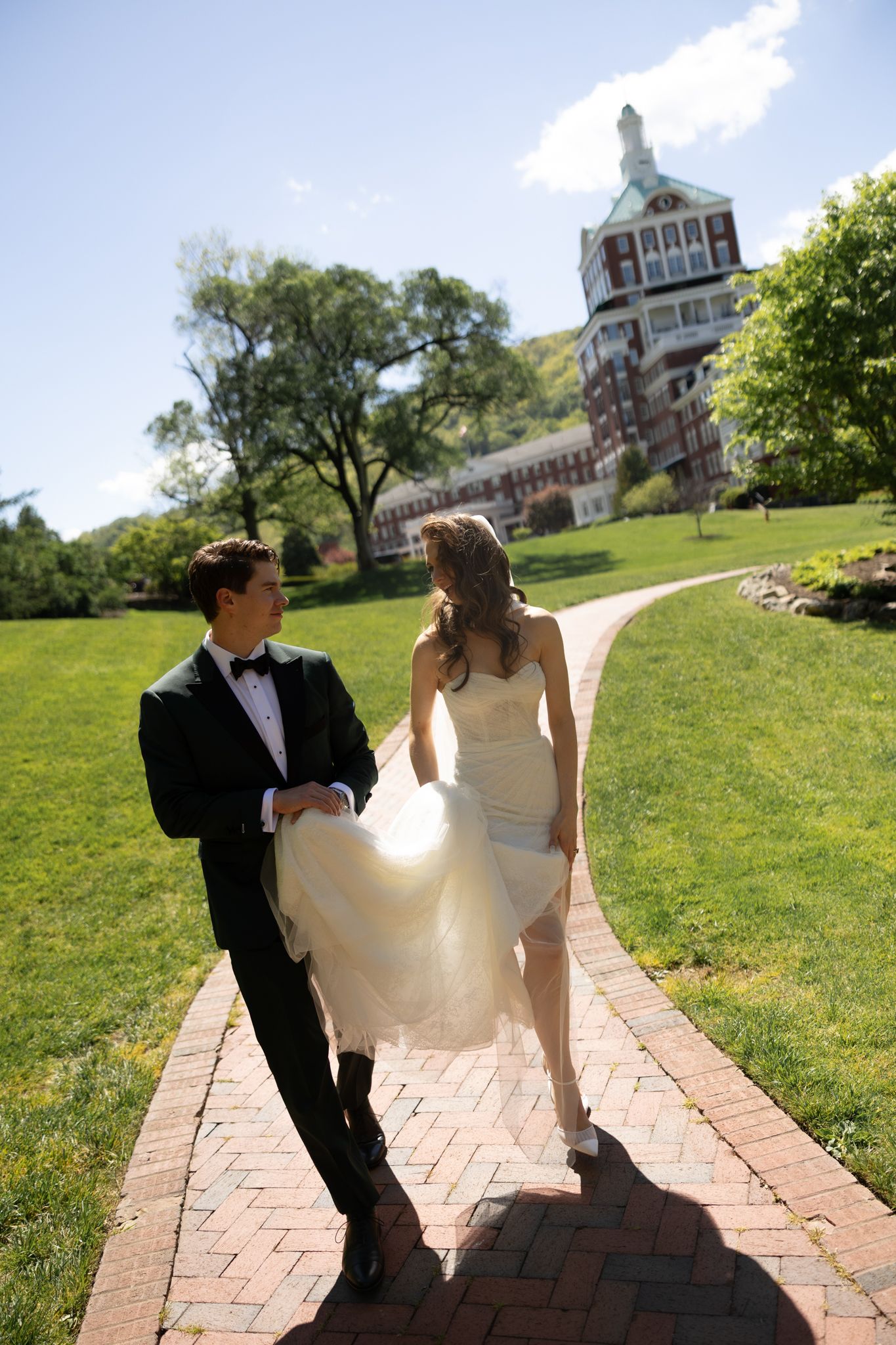 Bride and groom walking hand-in-hand on a brick path with a grand estate building and green lawn in the background.