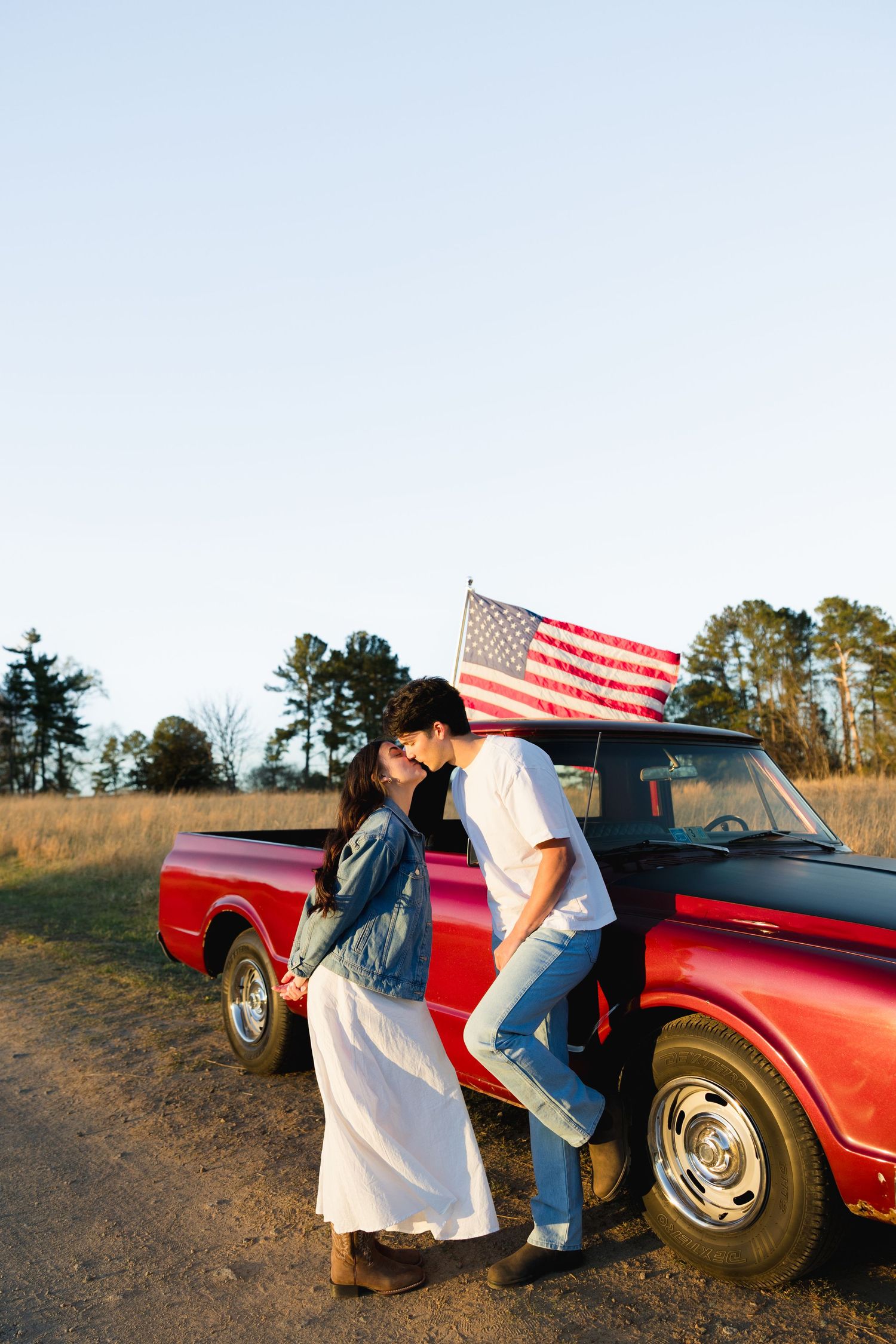 A romantic couple sharing a kiss beside a red vintage truck with an American flag in a rural field at sunset.
