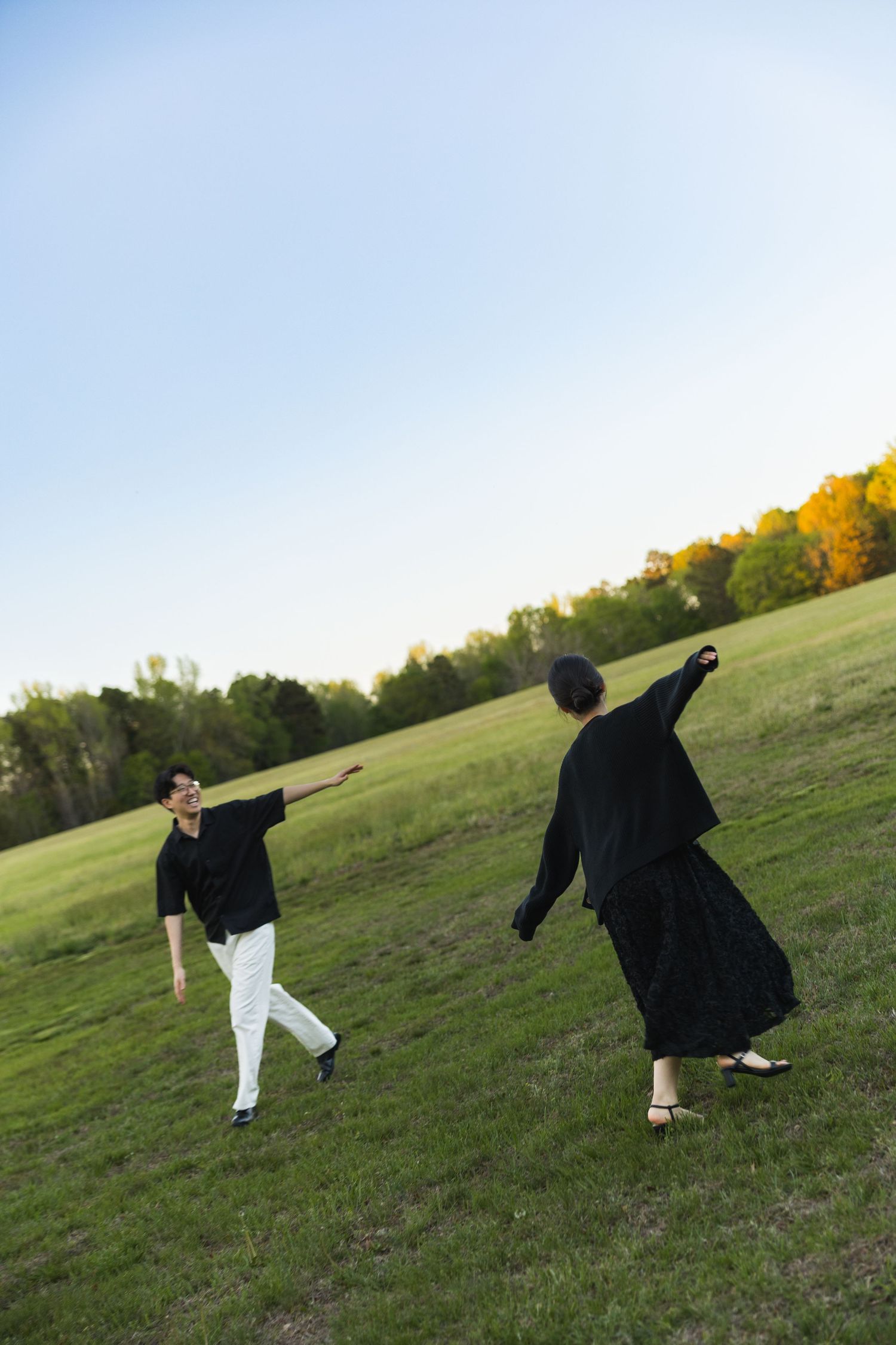 A couple playfully poses in an open green field at sunset, both dressed in black and white outfits.
