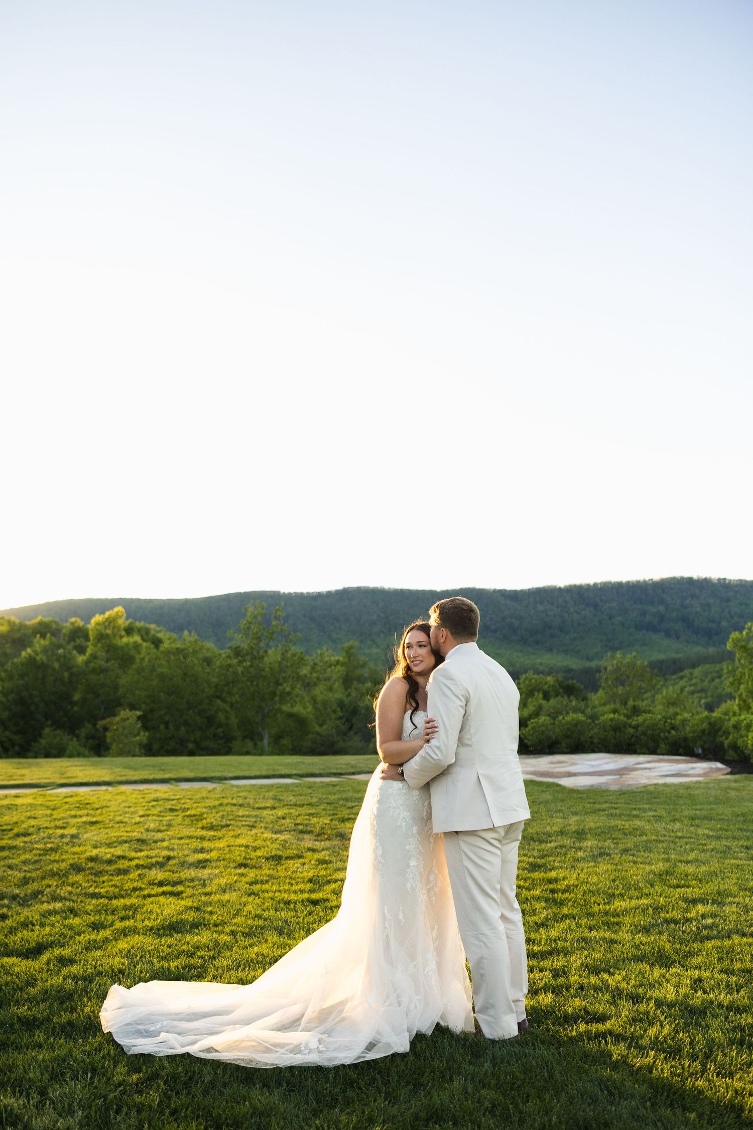 Bride and groom embrace on lush green lawn at golden hour with stunning mountain backdrop on their wedding day.