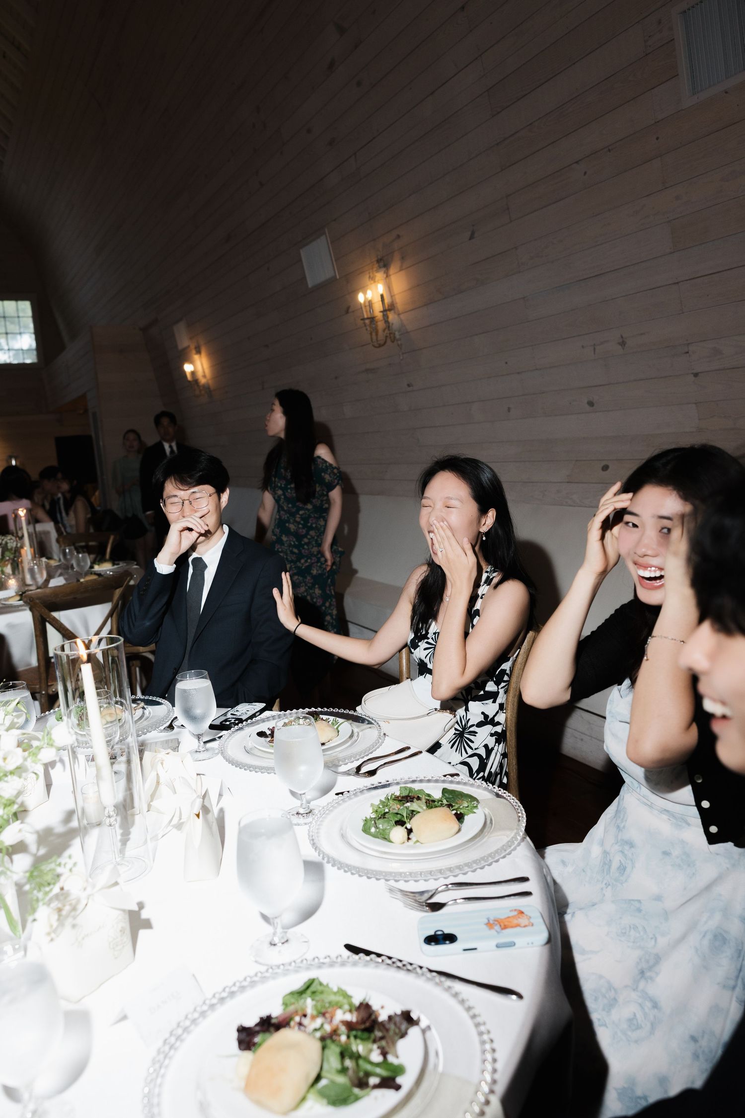 Guests react with surprise and laughter at a formal wedding reception dinner table with elegant place settings.