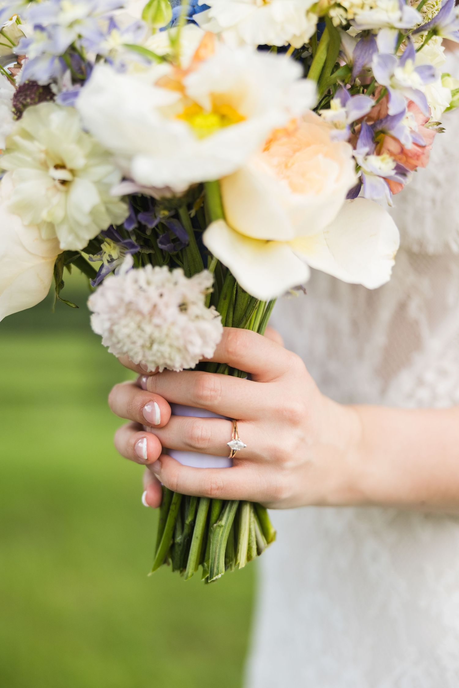 Bride holding a colorful wedding bouquet with white, peach, and purple flowers, wearing a diamond engagement ring.