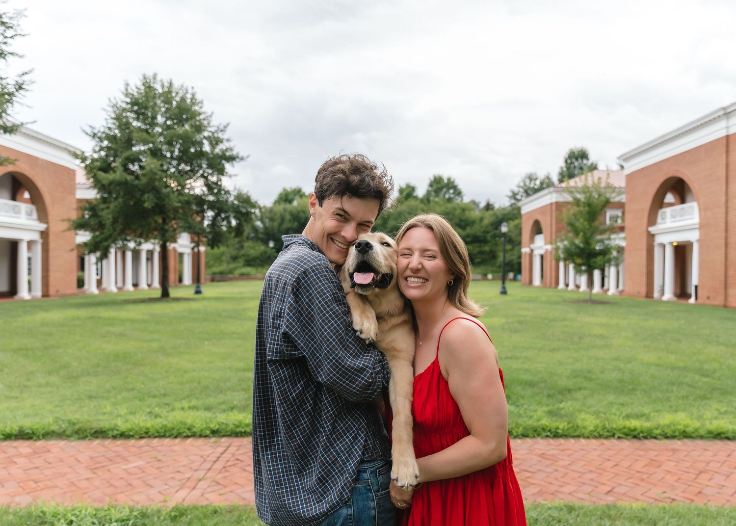 Happy couple holding an adorable dog in front of a brick building with arched walkways.