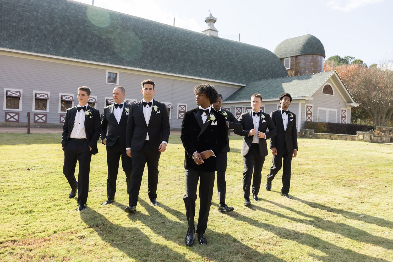 Groomsmen in black tuxedos walking outdoors near a rustic barn venue on a sunny wedding day.