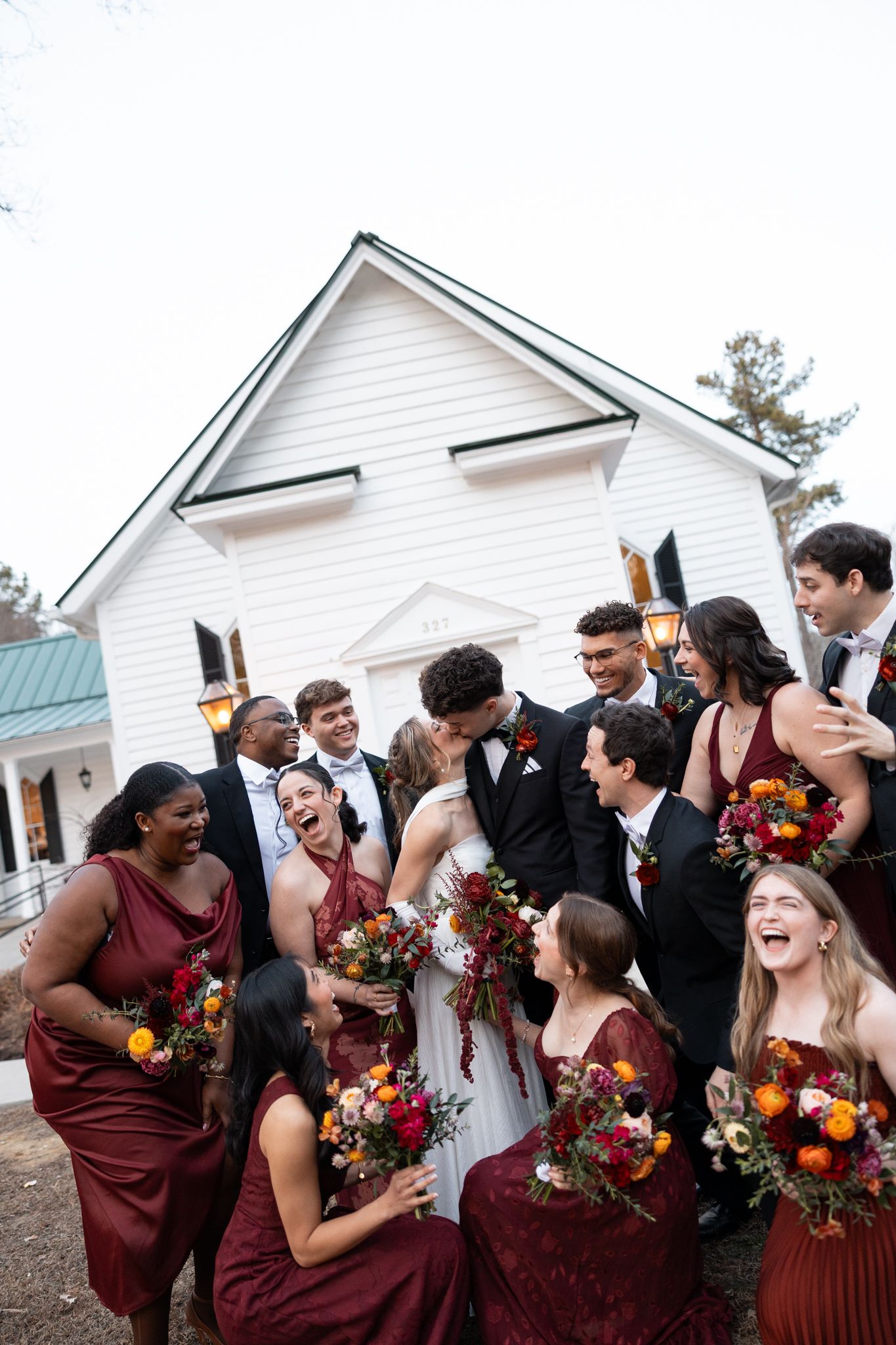 Wedding party in burgundy dresses and black tuxedos laughs together outside a white chapel venue.