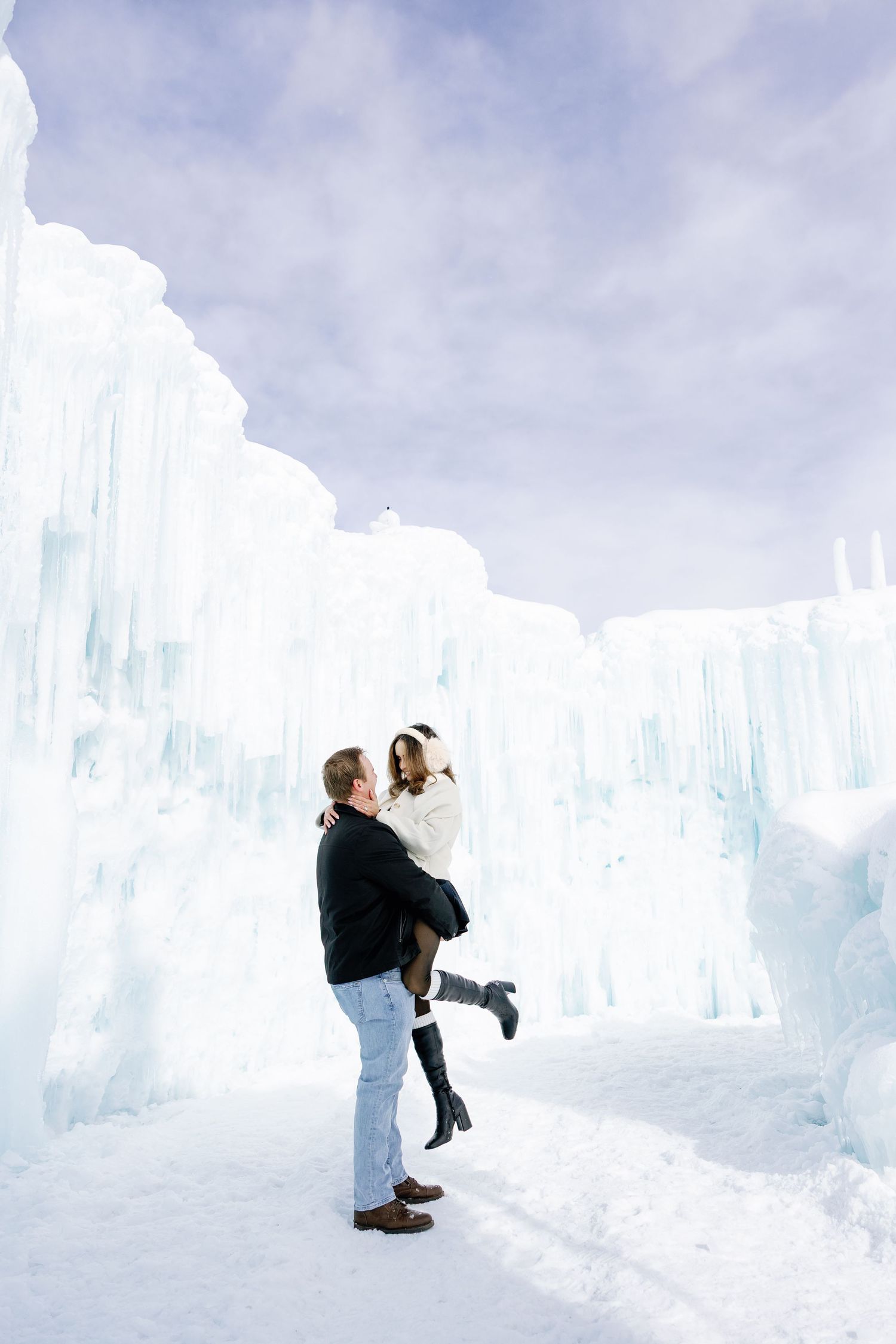 Man lifting woman in front of towering blue ice castle formations during a romantic winter engagement shoot.