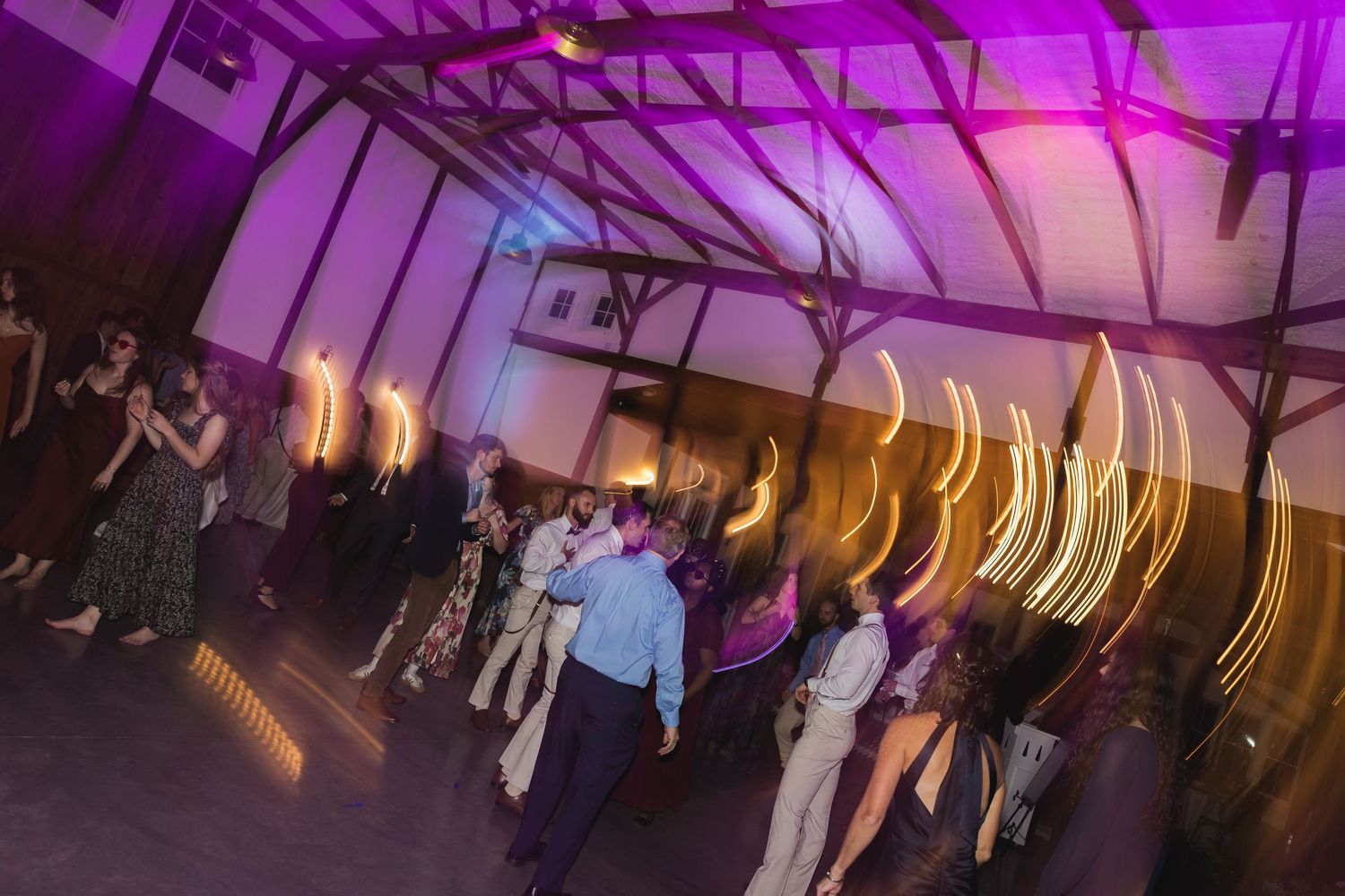 Wedding guests dancing under colorful purple lights at a lively rustic barn reception.