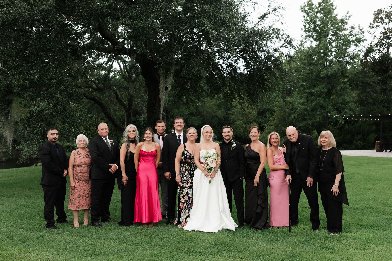 Wedding party poses outdoors on green lawn, bride in white gown holding bouquet, guests in pink and black formal attire.