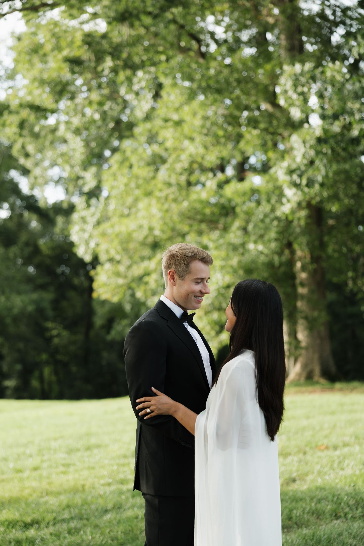 A smiling groom in a black tuxedo gazes at his bride in a white cape gown outdoors in a lush green park.