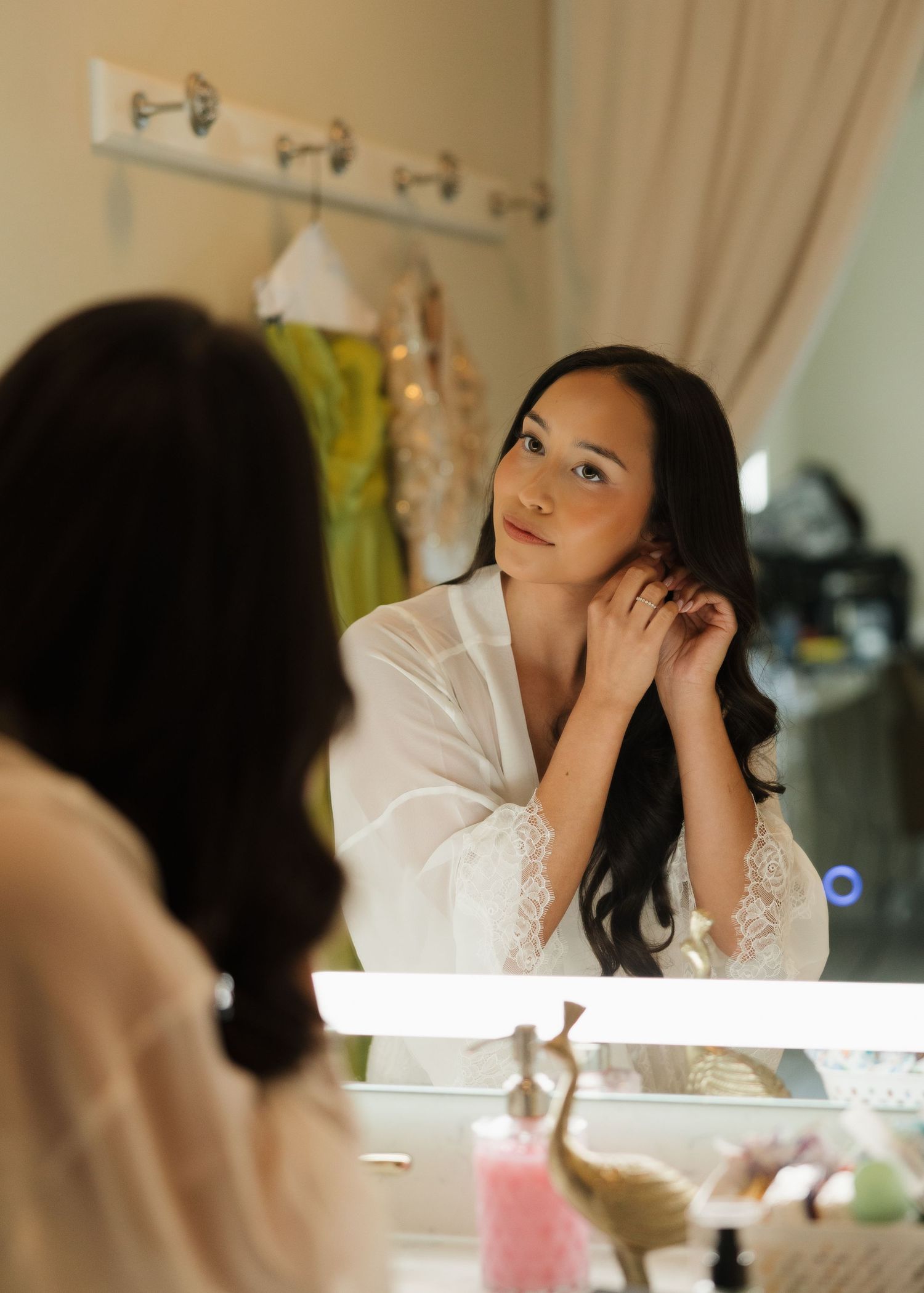 A bride in a white lace robe adjusts her earring while gazing into a lighted vanity mirror while getting ready.