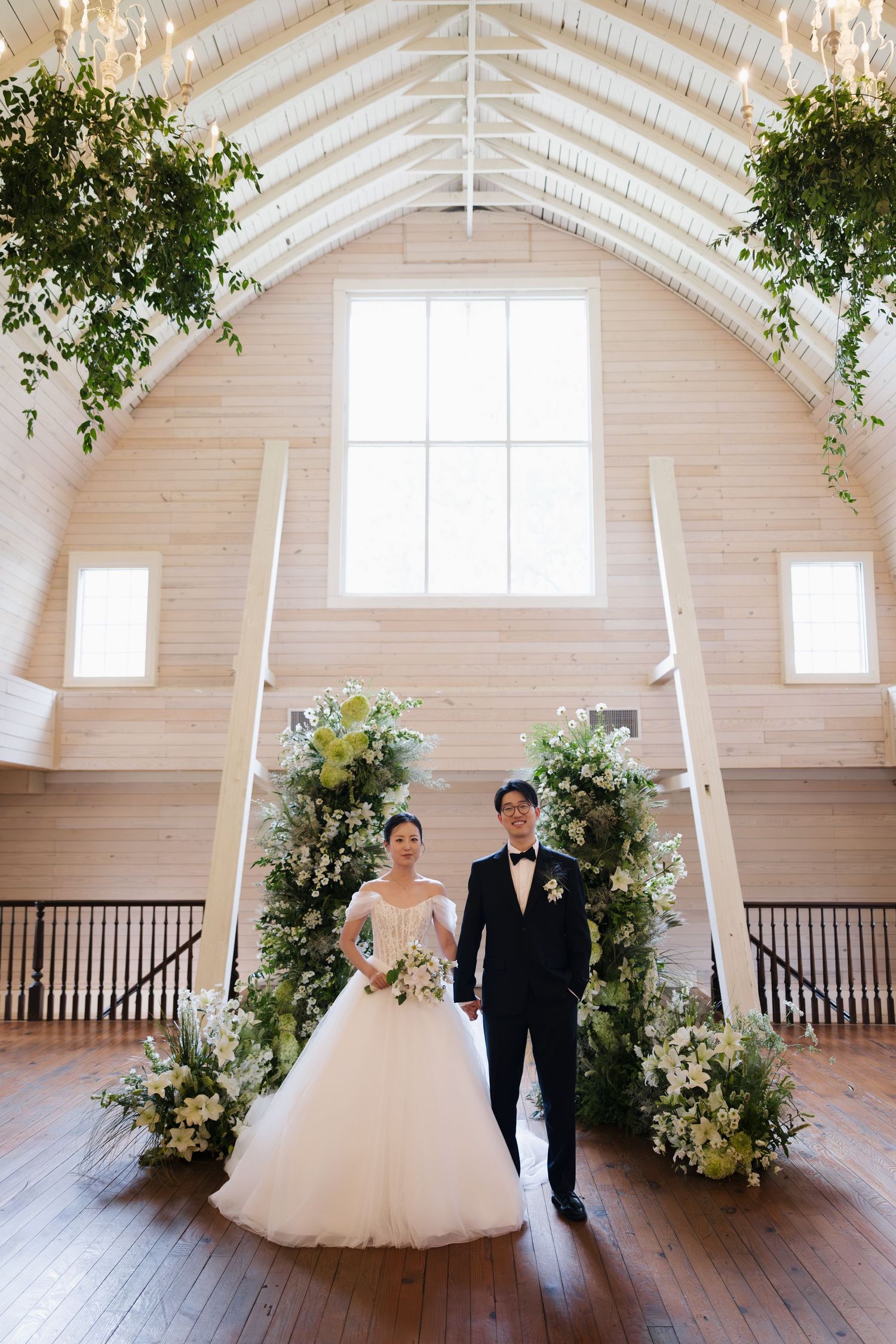 Bride and groom stand beneath floral arch in elegant barn wedding venue with vaulted wood ceiling and lush greenery.