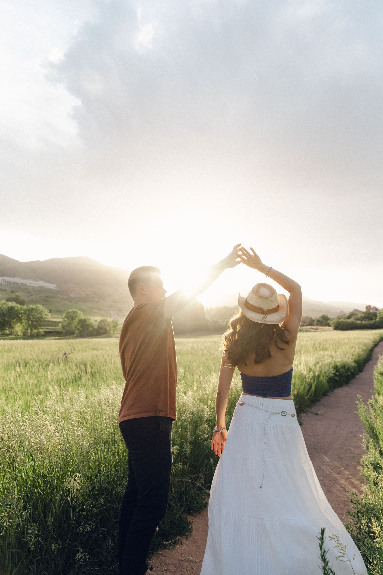 Couple dancing on a dirt path through green fields with mountains and golden sunset behind them.