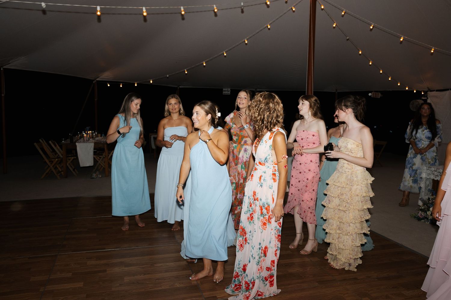 Group of women in pastel dresses gathered on dance floor under string lights at wedding reception.