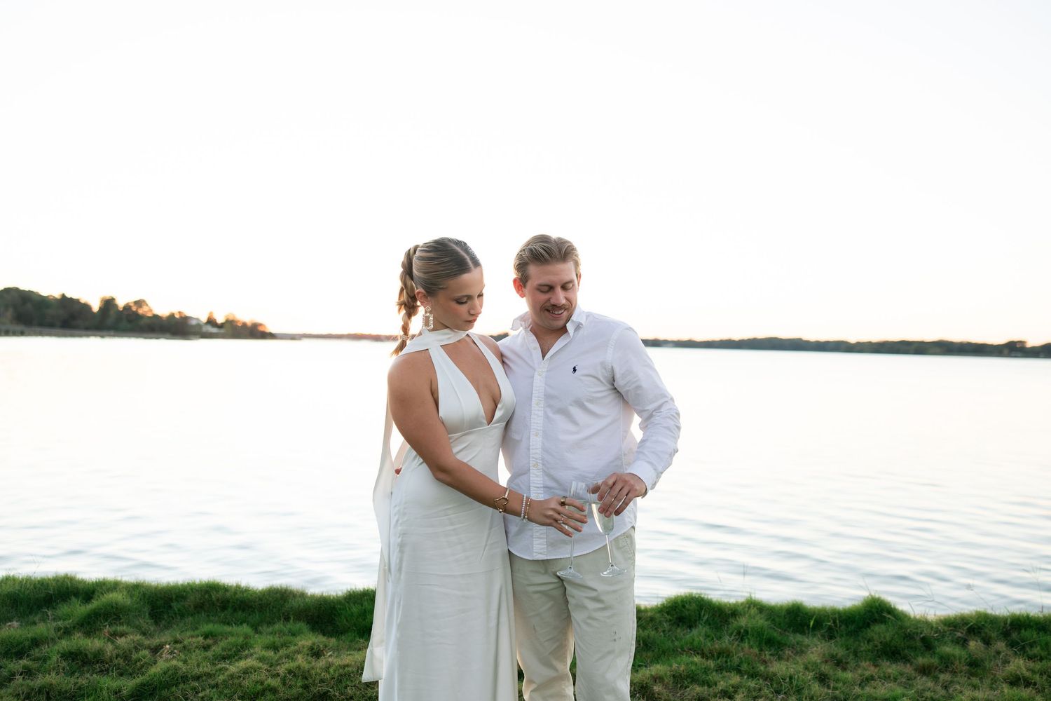 Couple in white outfits standing by a serene lake at sunset, man tenderly looking at woman's baby bump.