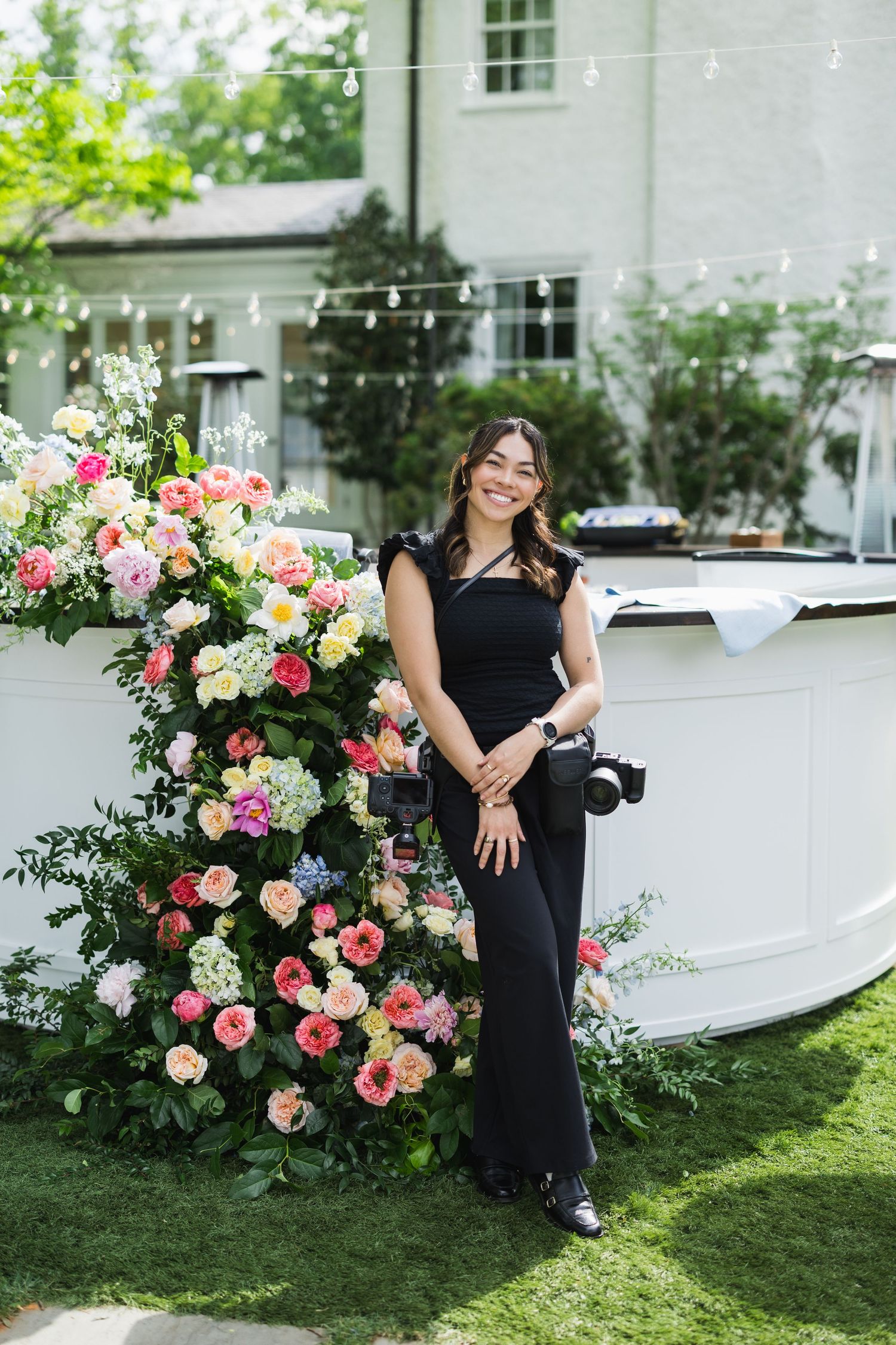Woman in black outfit posing beside colorful floral arrangement at outdoor event with string lights.