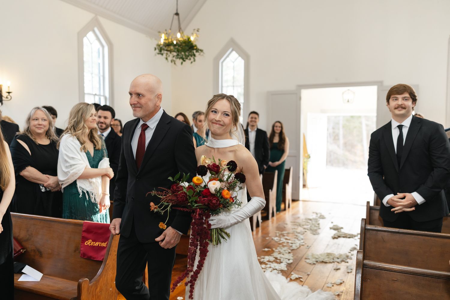 Smiling bride in white gown carries colorful bouquet while walking down church aisle with her father.