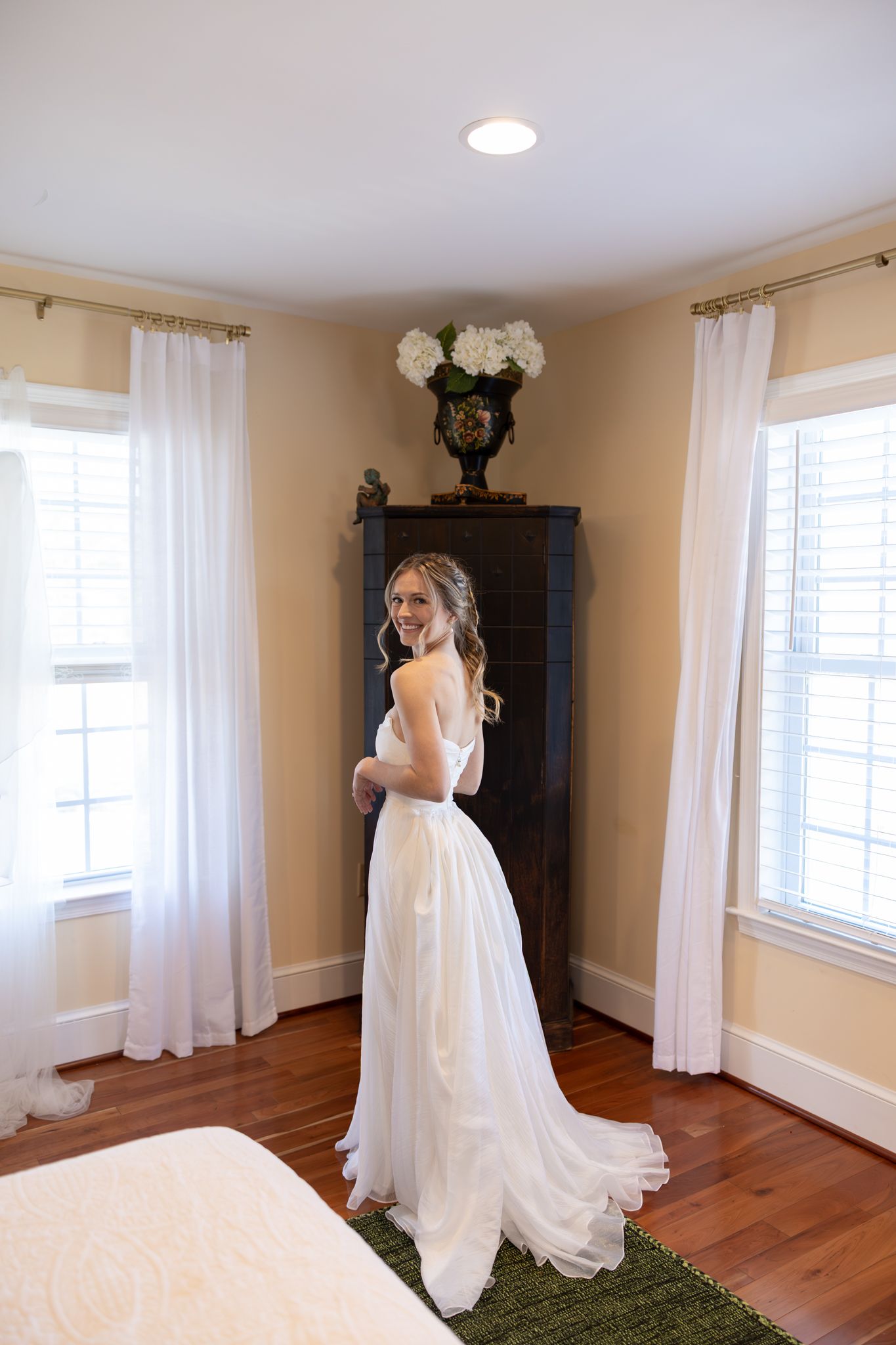 Bride in a flowing white wedding gown standing in a sunlit bedroom looking over her shoulder.