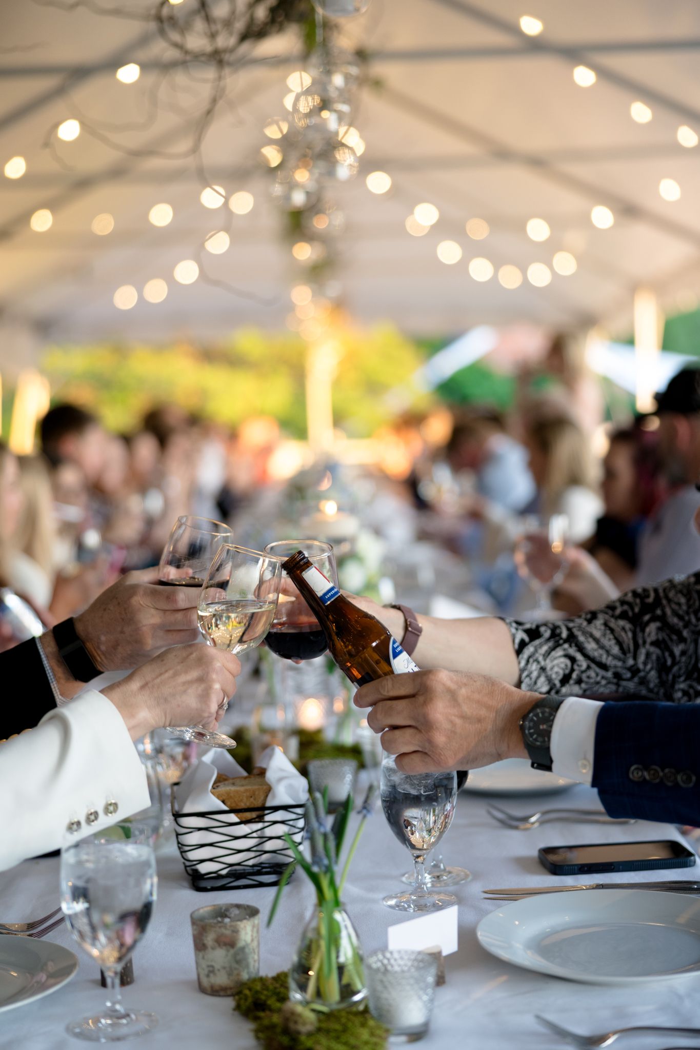 Guests clink glasses and pour wine at an elegant rehearsal dinner table under a white tent with string lights.