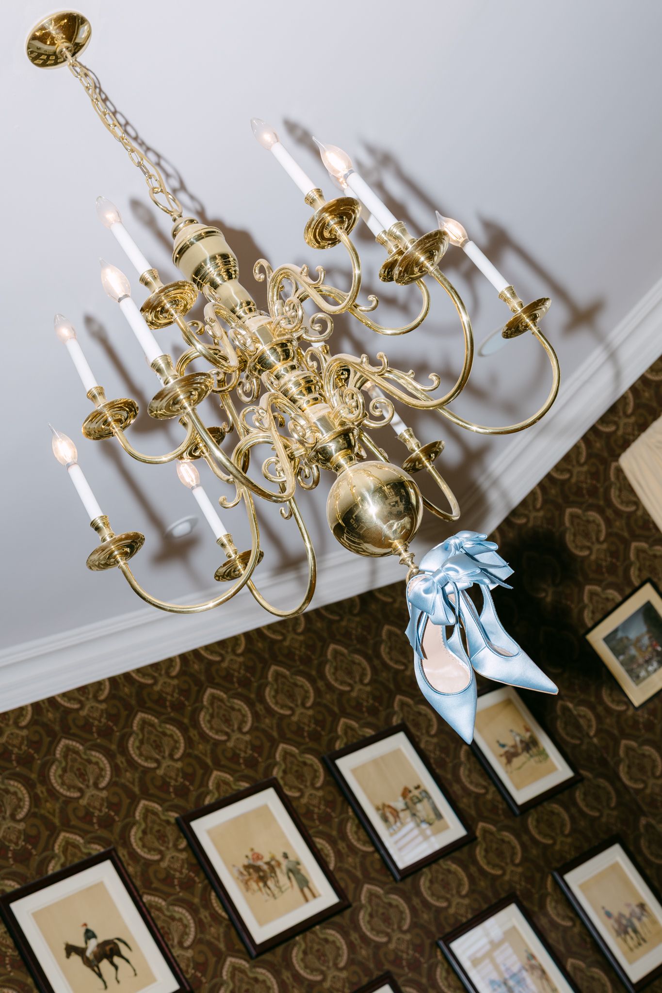 Light blue satin bow heels hanging from ornate brass candelabra chandelier above framed equestrian artwork.