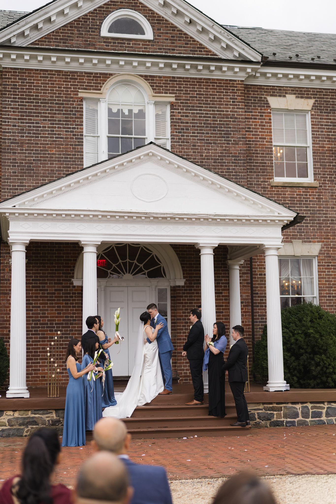 Wedding couple kissing on steps of elegant brick colonial venue surrounded by bridesmaids in blue dresses and groomsmen.