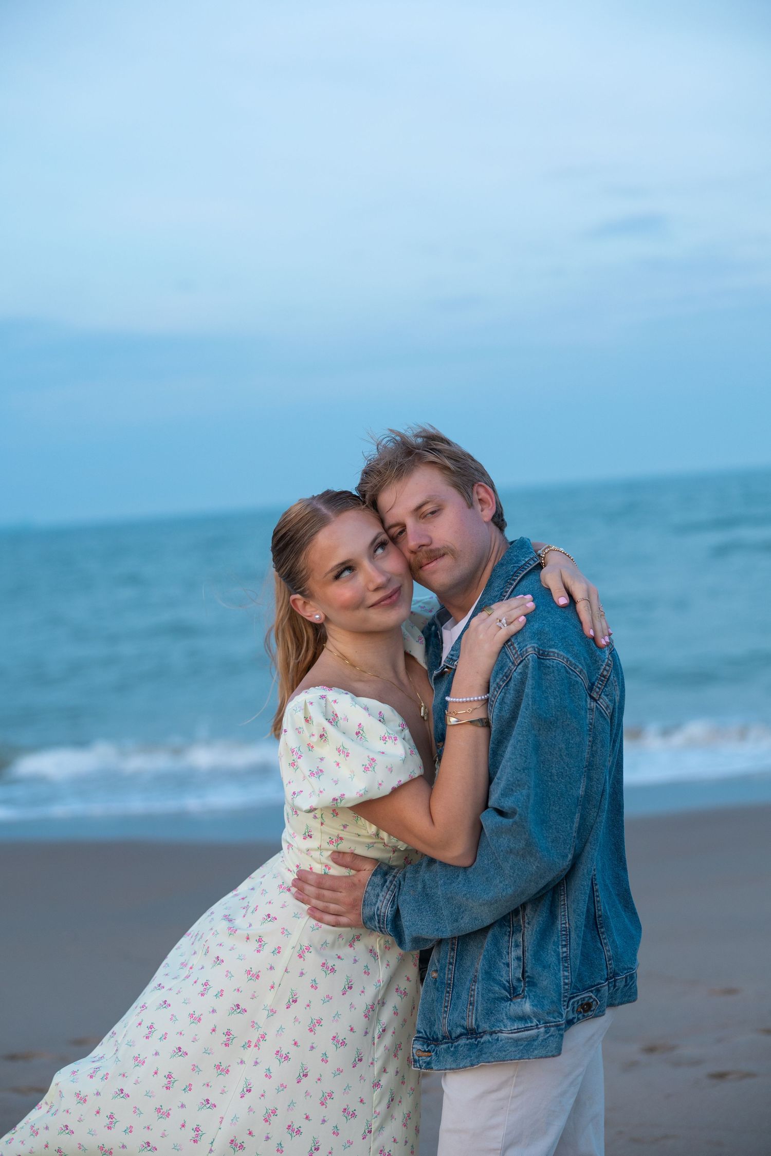Loving couple embracing on beach at dusk, woman in floral dress and man in denim jacket.