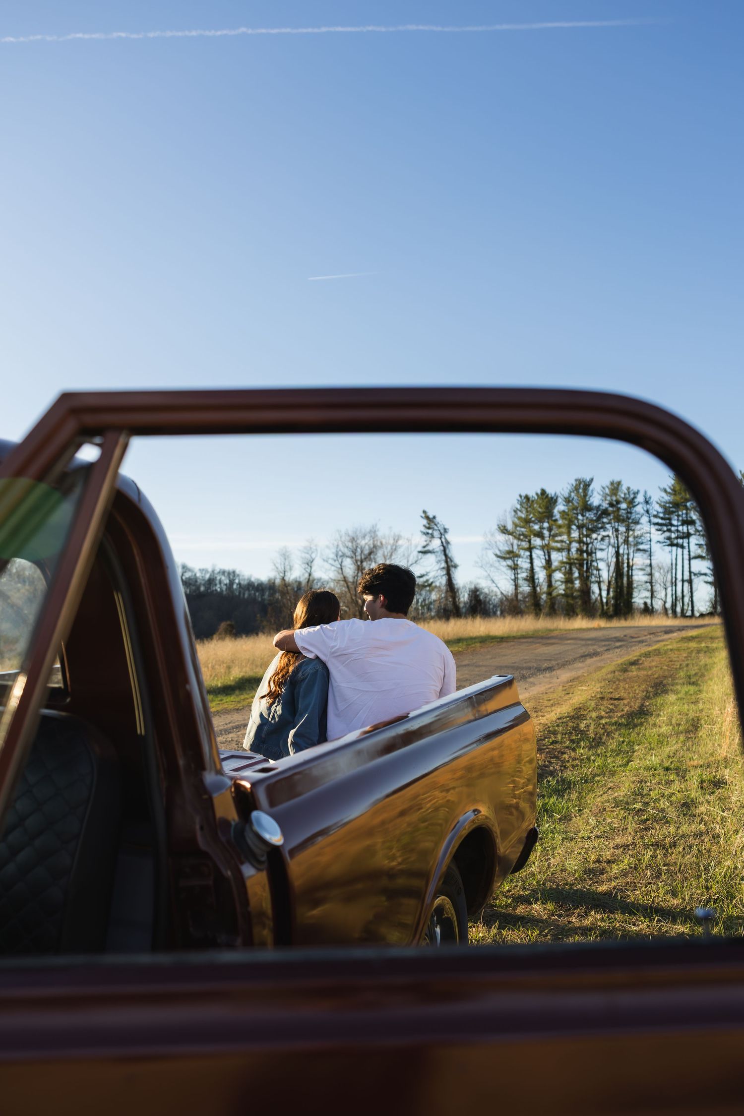Couple sitting on truck tailgate viewed through vintage red pickup window, overlooking scenic field.