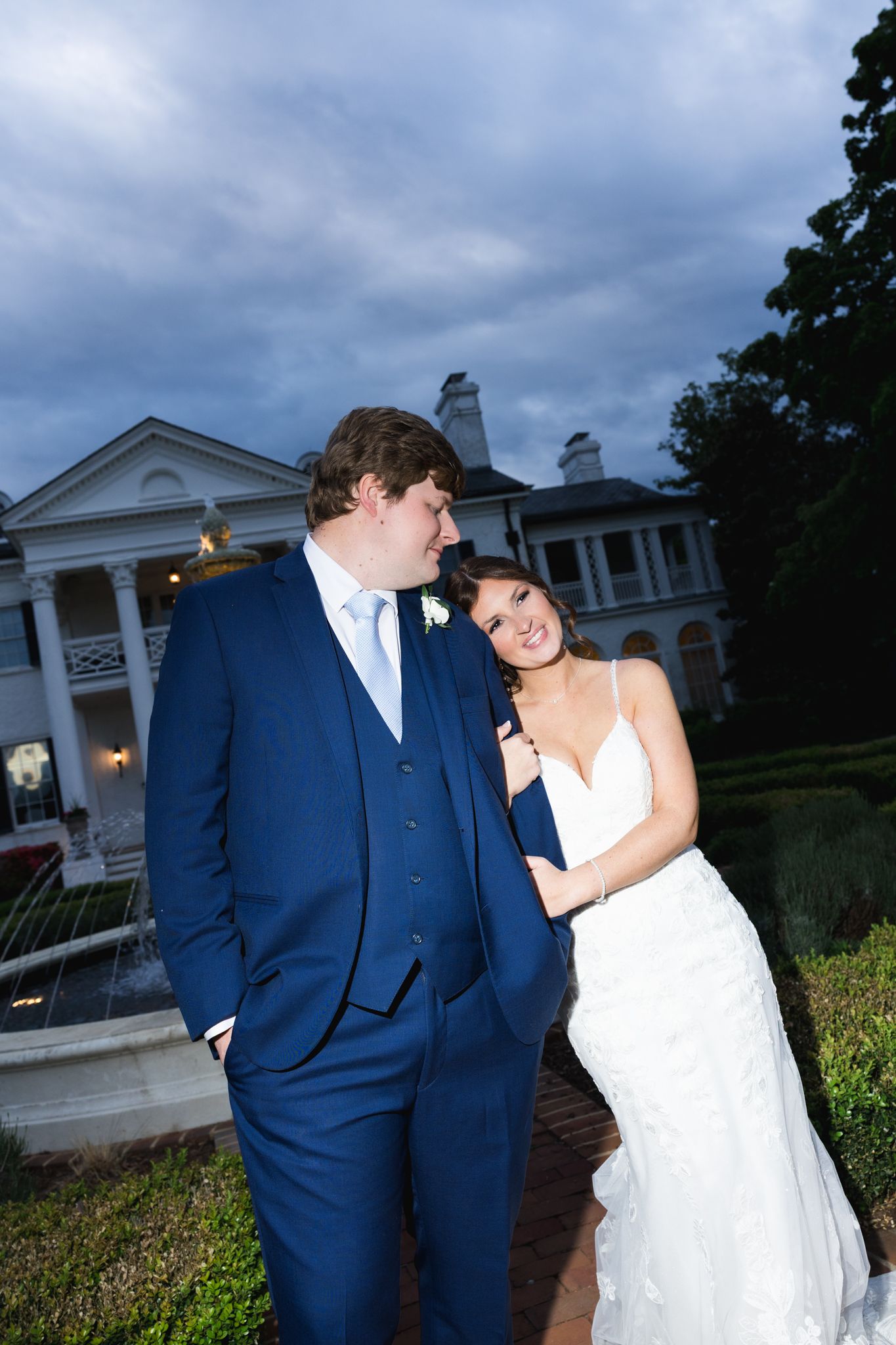 Bride and groom posing outside elegant mansion at dusk, groom in navy suit, bride in white lace gown.