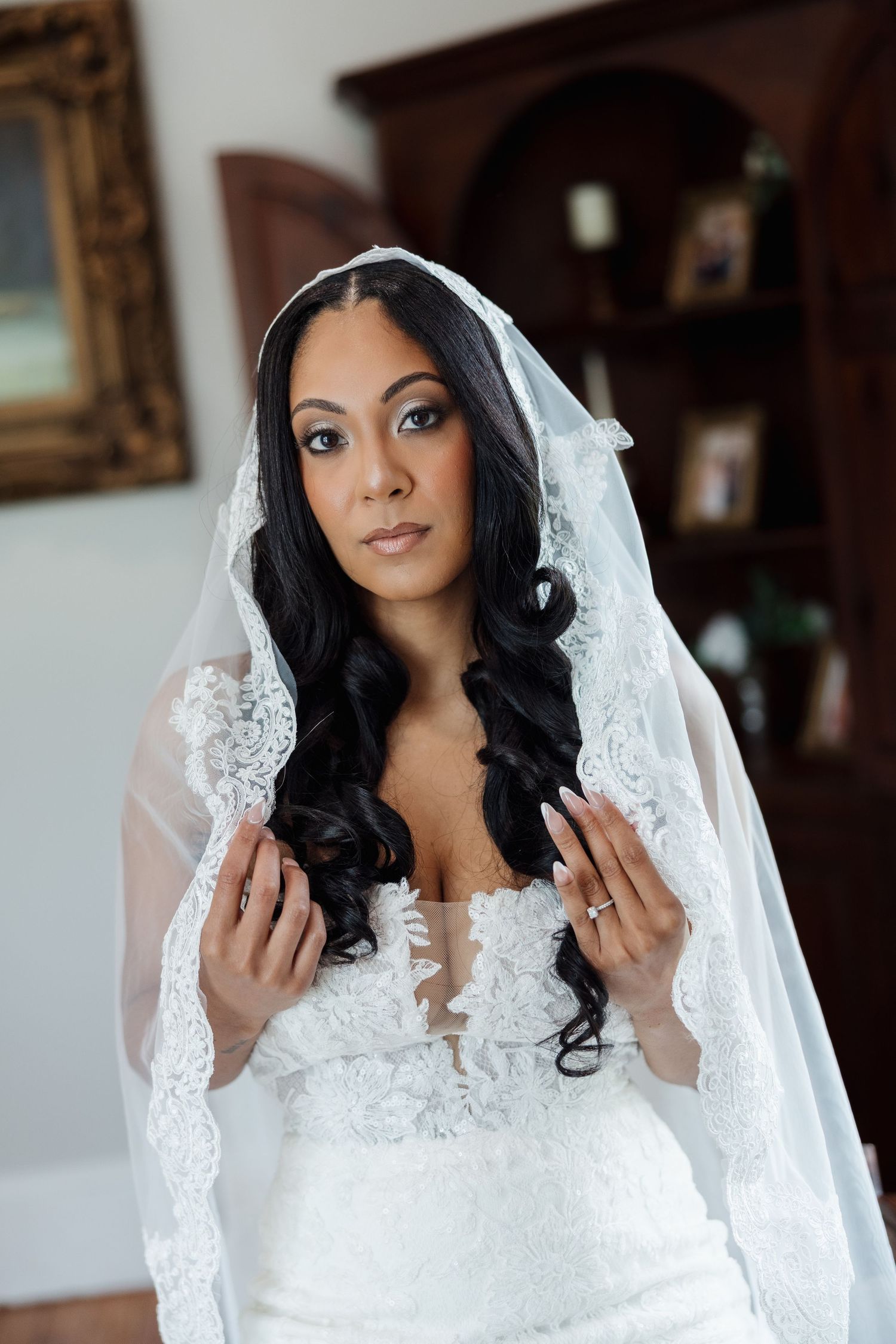 Bride in white lace wedding dress and veil with long dark curly hair posing indoors before her wedding ceremony.