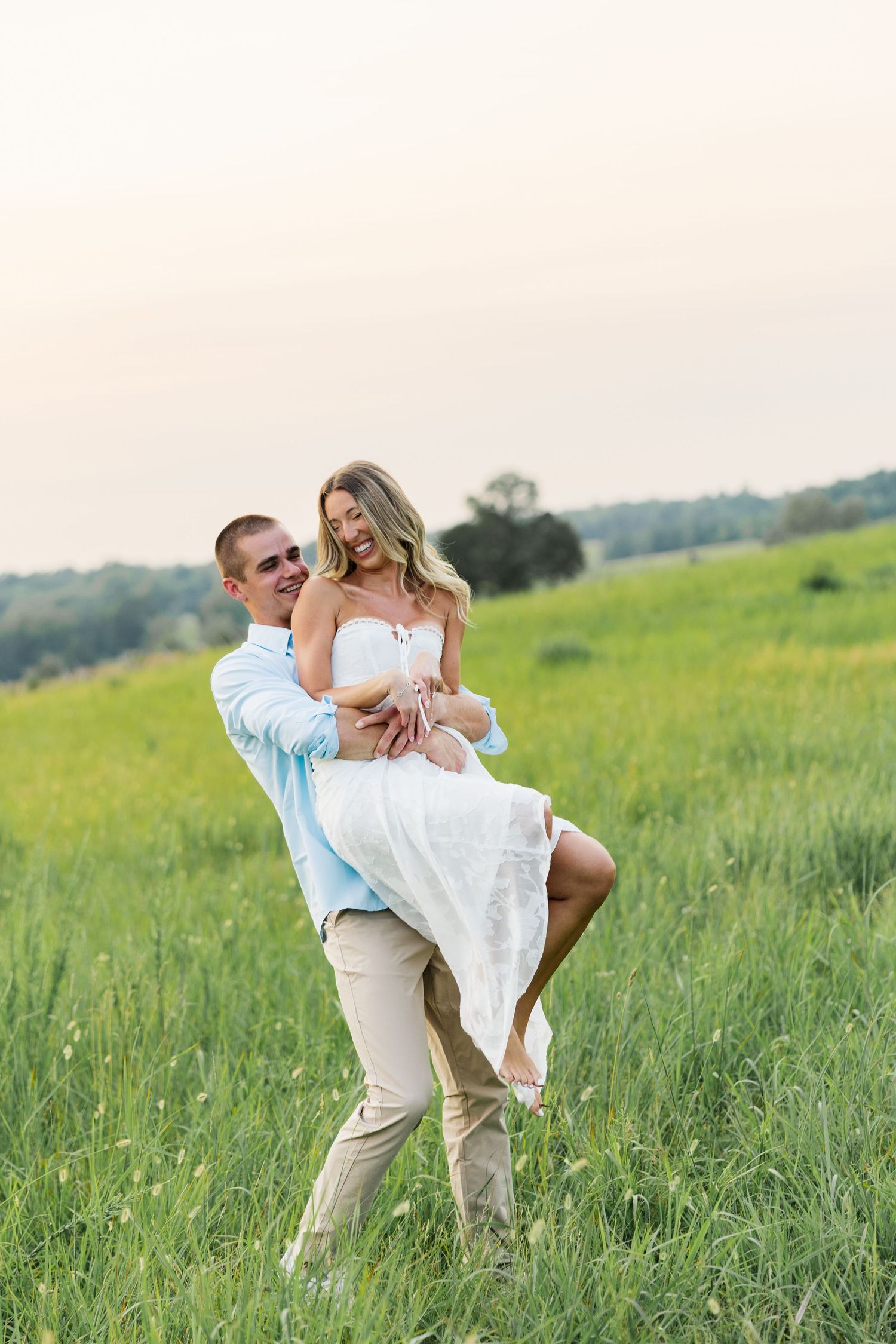 A man lifts a smiling woman in a white dress in a lush green field during a romantic outdoor engagement session.