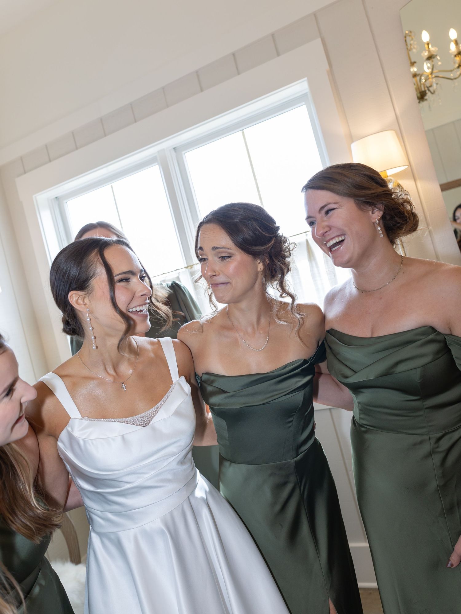 Bride laughing joyfully with bridesmaids in dark green gowns while getting ready in a bright white room.