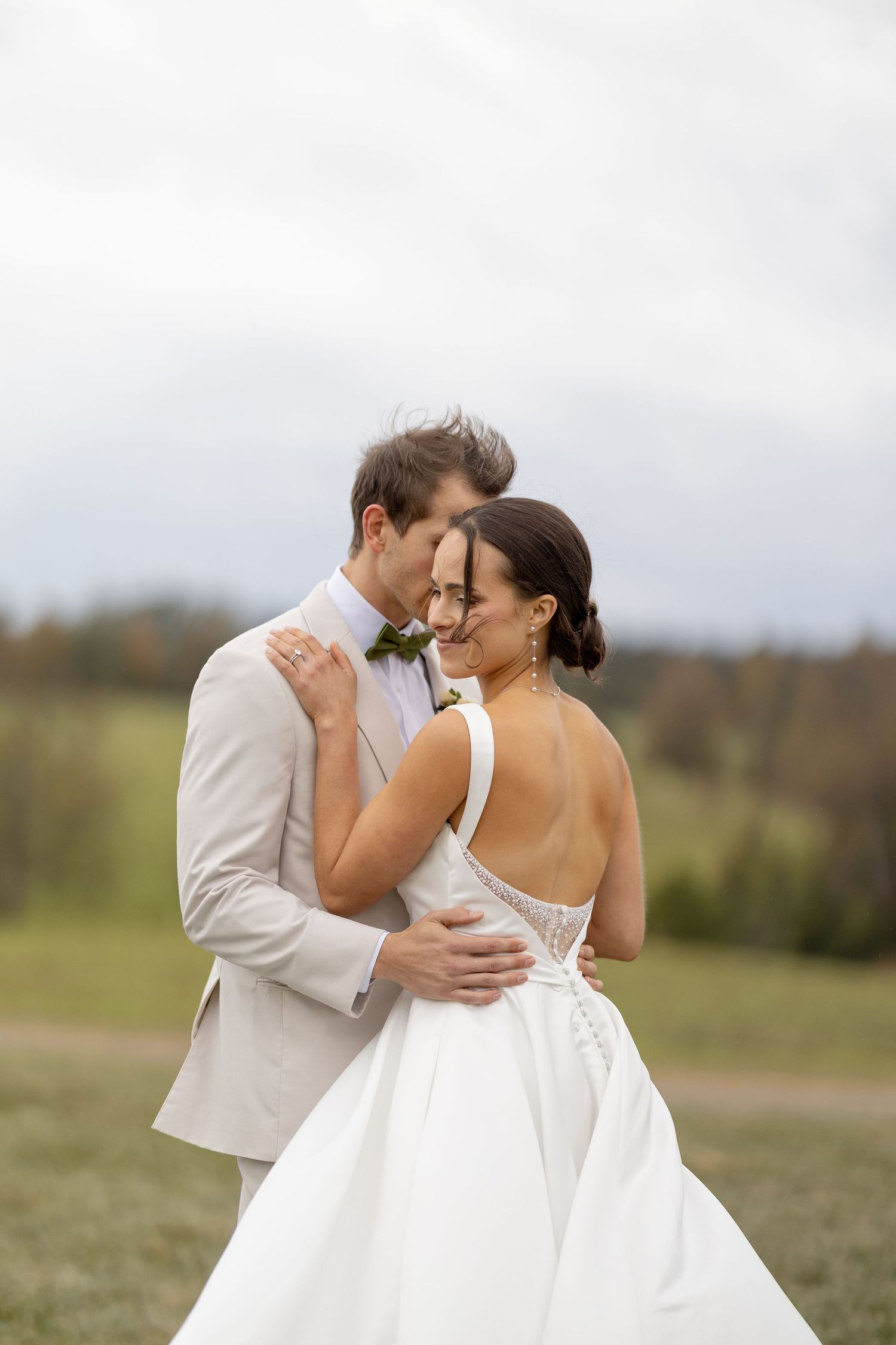 Groom in ivory suit embraces bride in backless white gown in a romantic outdoor field portrait.